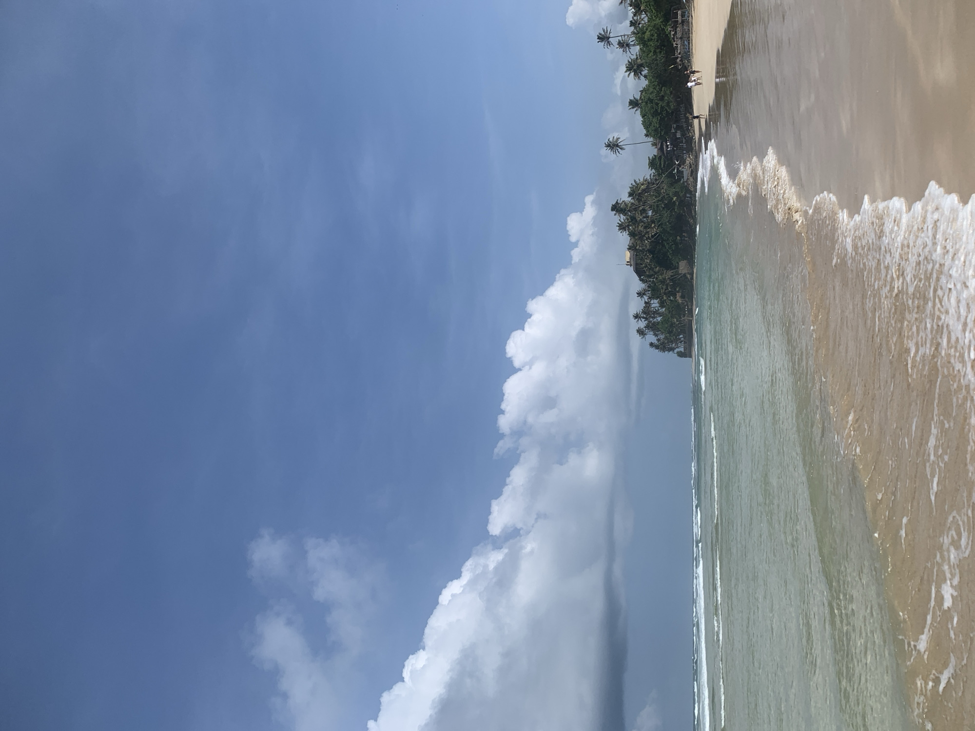 The image depicts a tropical beach scene in Kathaluwa, Sri Lanka, on a clear day with scattered clouds. The foreground is dominated by a sandy beach with gentle waves lapping at the shore. The water is a clear, light green, and the sand is a warm, light brown. In the midground, a line of palm trees and lush green vegetation stretches along the coastline, suggesting a tropical island setting. Some structures, possibly small buildings or huts, are visible amongst the trees. A few people are walking along the beach in the distance, appearing as small figures against the vastness of the landscape. The sky is a bright blue with a few large, white, fluffy clouds, particularly on the left side of the frame. The overall impression is one of a serene and picturesque coastal environment.