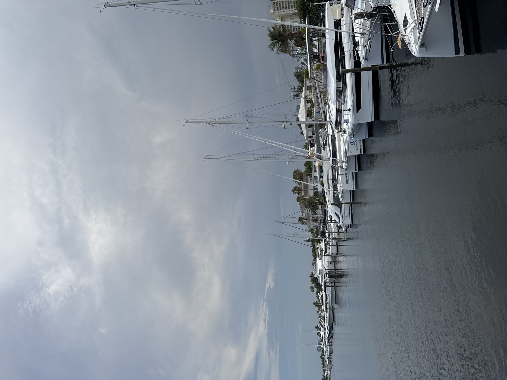 The image depicts a harbor in Fort Lauderdale, Florida, USA, on a cloudy day. Numerous sailboats and yachts are docked along a waterfront. The boats are primarily white, with visible masts and rigging. In the background, buildings, palm trees, and a hazy sky are visible. The water is dark and reflective, showing ripples from the boats. No people are visible in the frame.