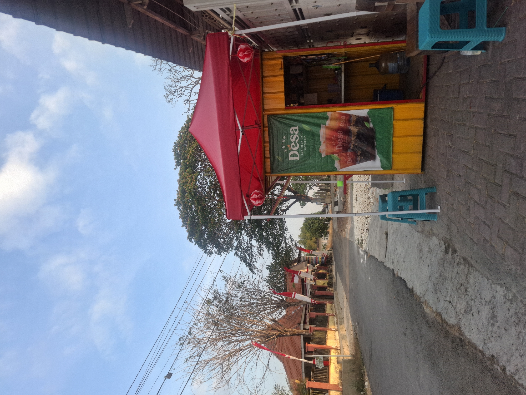 This is an eye-level, medium shot of a small, roadside food stall with a red canopy in Simbatan, Indonesia. The stall is yellow and features a green banner advertising "Esteh Desa" with images of iced tea. Two turquoise plastic stools are positioned in front of the stall, and a larger blue plastic bin is visible inside. To the left of the stall, a paved road runs past a building adorned with red and white Indonesian flags. Bare trees and a large, leafy tree are also present in the background. The sky is clear with a few wispy clouds. The scene appears to be a quiet, early morning setting.