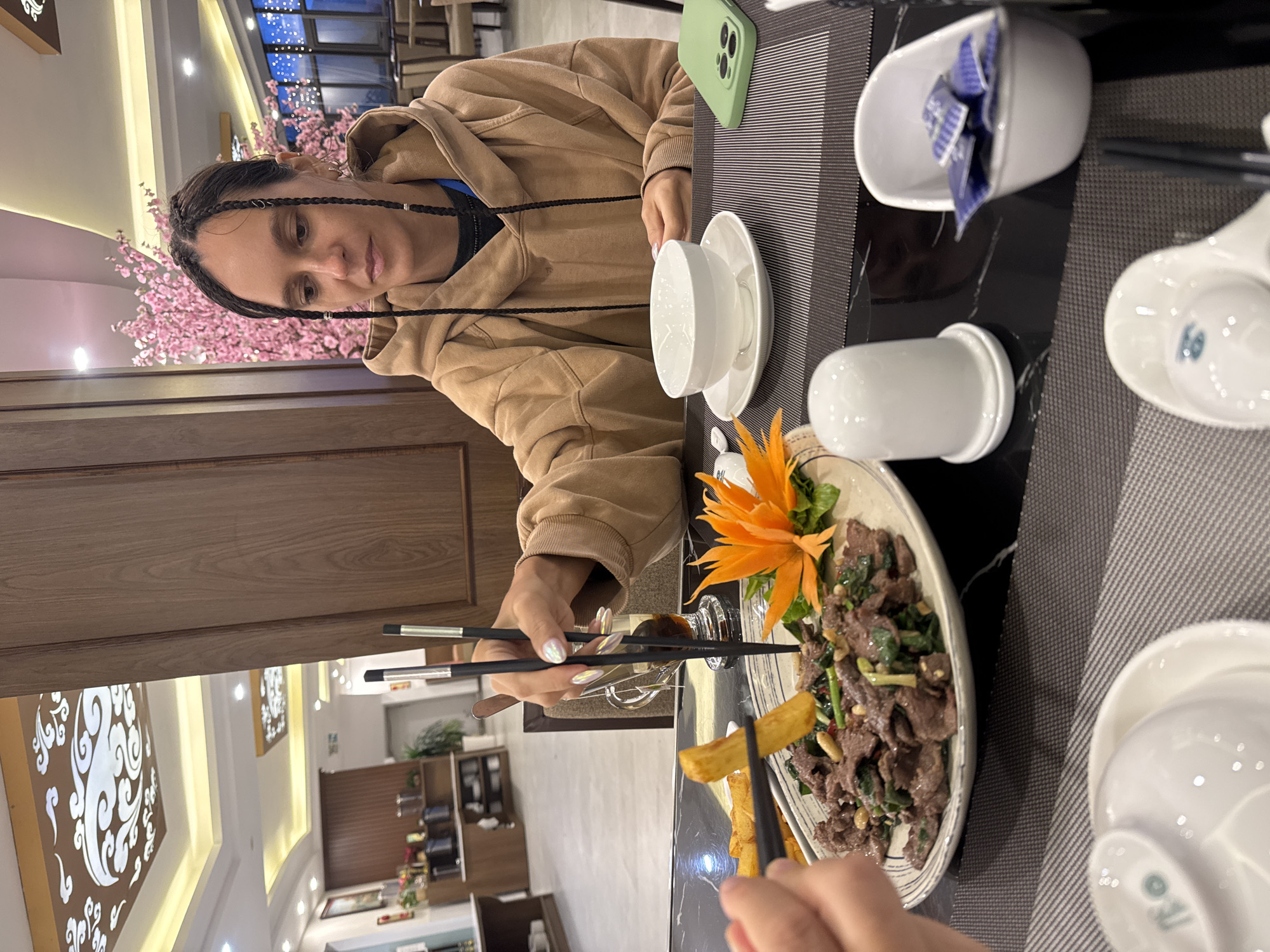 A woman with braided hair is seated at a restaurant table in Binh Lu, Vietnam. She is wearing a tan hoodie. With chopsticks, she is picking up a french fry from a plate of meat and vegetables garnished with an orange flower. The table is set with white ceramic dishes, and a green cell phone rests on the striped section of the tabletop. In the background, a decorated wall and restaurant interior are visible.