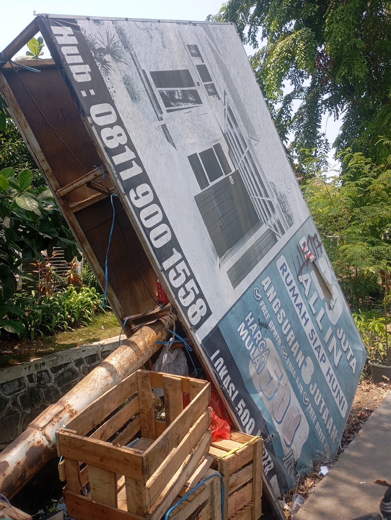 In Babelan, Indonesia, a large, damaged billboard depicting a house is propped up on a trailer. The billboard is tilted at a steep angle, with a portion of its surface torn and spray-painted with graffiti. Text on the billboard includes "Hub: 0811 900 1558", "RUMAH SIAP HUNI", "ANGSURAN JUJTAN", and "HARGA MULAI LOKASI 500 JUTA". A wooden crate filled with debris sits in front of the trailer, and a large, rusty metal pipe extends from the trailer towards the foreground. The scene is set outdoors, with lush green foliage and trees visible in the background, suggesting a sunny day. The ground appears to be a mixture of dirt and pavement. No people are present in the image.