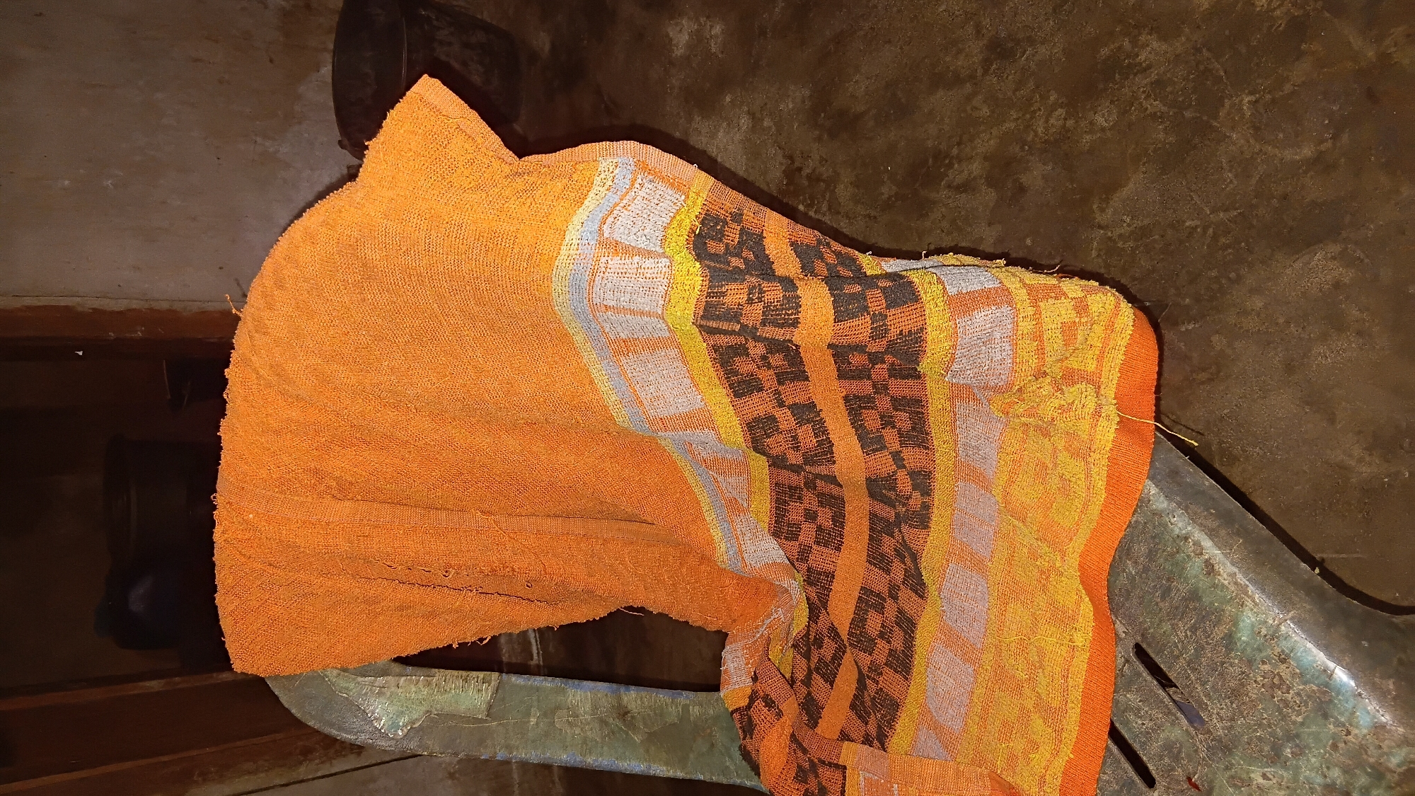 An orange patterned textile is draped over the back of a weathered, light-colored plastic chair. The textile has a woven pattern with horizontal bands of grey, yellow, and dark brown geometric shapes. The chair is situated in an indoor setting with a concrete floor and a wall in the background. The lighting appears to be artificial, creating shadows and highlighting the textures of the objects. The overall impression is one of a simple, utilitarian space. This image does not contain any specific details that would allow for the determination of its location within Prabumulih, Indonesia.