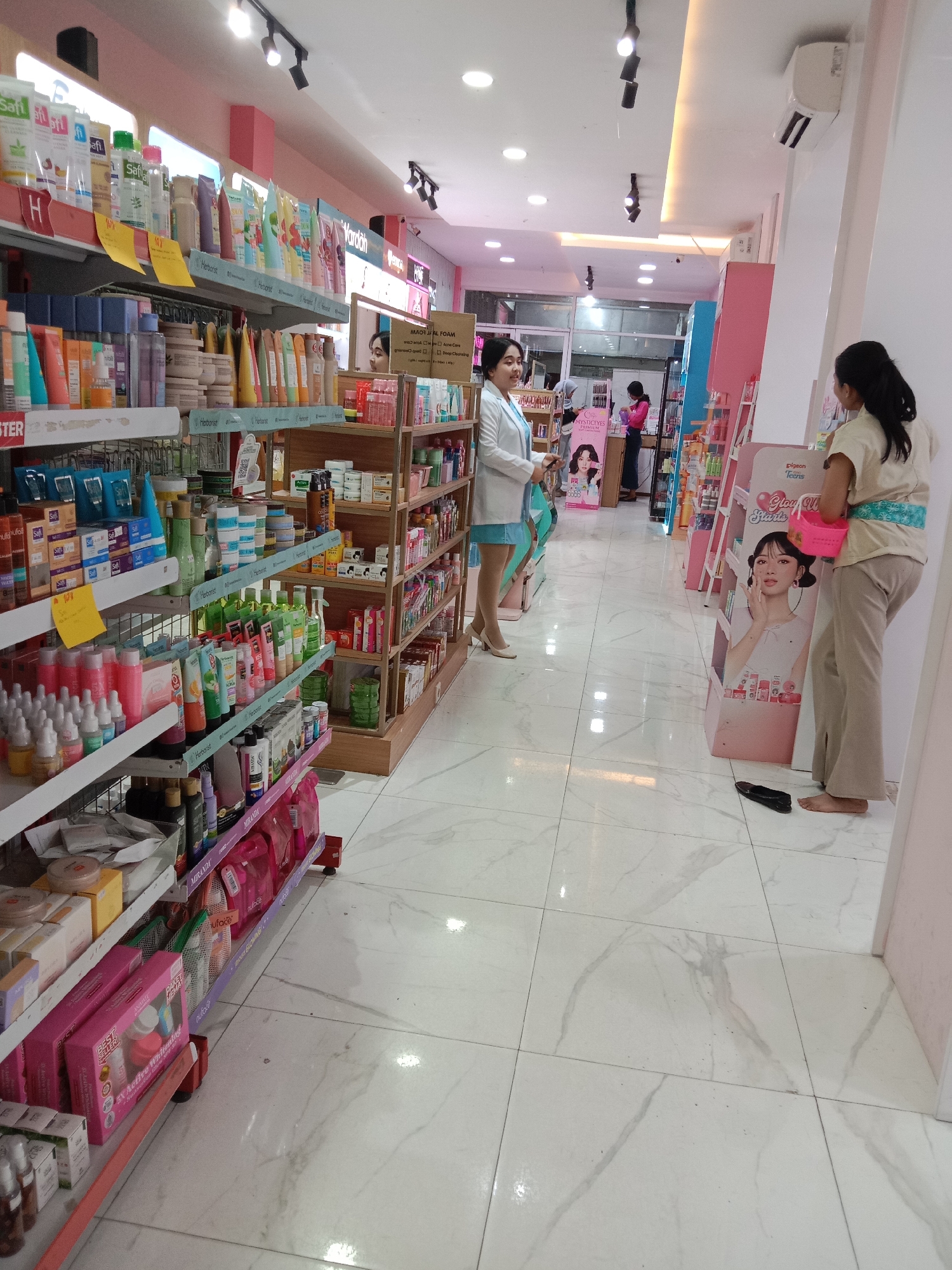 The image depicts the interior of a cosmetic store in Kaliwungu, Indonesia. The store is well-lit, with bright overhead lights and spotlights. Rows of shelves are stocked with various cosmetic products, including skincare items, makeup, and personal care products. The shelves are organized and neatly arranged, with products displayed in an appealing manner.
In the foreground, a woman in a light-colored top and beige pants is standing on the right side of the image, facing away from the camera. She is holding a pink basket. To her left, a woman in a white lab coat over a blue dress is standing near a display of products. She is looking towards the left and appears to be interacting with a customer or restocking shelves. Further in the background, another woman is visible behind a counter, also appearing to be an employee. A large poster of a woman's face is displayed prominently on a stand to the right of the central aisle.
The floor is tiled with a light-colored marble pattern. The overall atmosphere of the store is clean, organized, and inviting, suggesting a typical retail environment for cosmetic products.