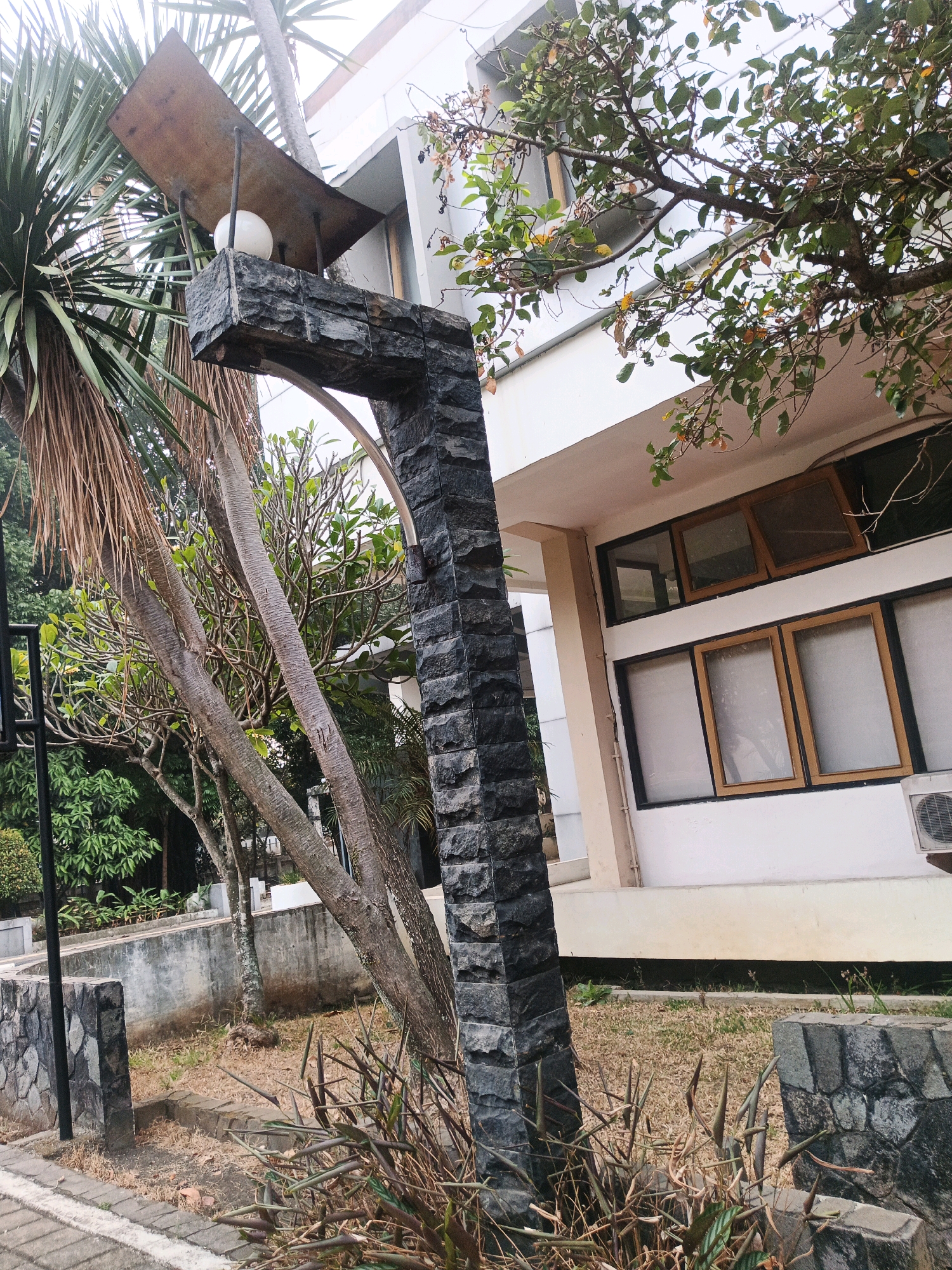 The image shows an outdoor scene with a building and vegetation. In the foreground, a stone pillar with an arched top is prominent. A white spherical light fixture is mounted on top of the pillar, under a metal canopy. Surrounding the pillar are dry grass, some brown leafy plants, and the trunks of two palm-like trees. In the background, a white building with several windows is visible. The windows have wooden frames and appear to be either tinted or frosted. An air conditioning unit is mounted on the wall of the building. The overall environment suggests a tropical or subtropical climate, possibly in a public space or garden area. The scene is static and does not depict any people or animals. The lighting suggests it is daytime.