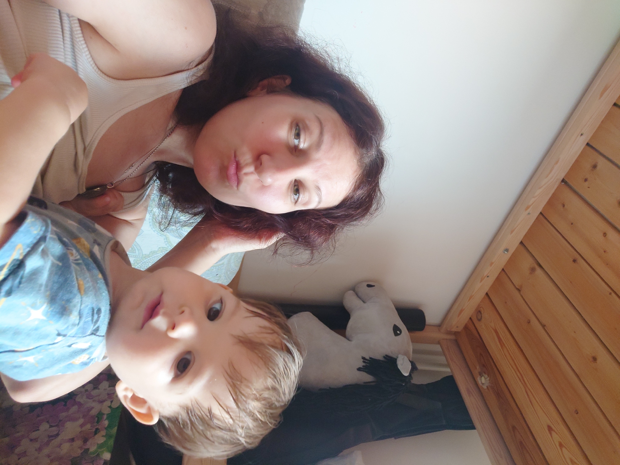 This is a selfie of a woman and a young child, likely mother and son, taken indoors. The woman, with dark hair and fair skin, is in the foreground, making a kissing face at the camera. She is wearing a beige tank top and a silver necklace. Next to her, the child, with fair hair and brown eyes, looks directly at the camera with a neutral expression. He is wearing a blue t-shirt with a star pattern. In the background, a stuffed toy horse lies on a dark surface. The room appears to be a bedroom or living area, with wooden paneling visible on the right side of the frame, suggesting a slanted ceiling or wall typical of an attic space. The lighting is soft and natural, implying daytime. The overall mood of the image is casual and intimate, capturing a moment between the two individuals. No visible text is present in the image.