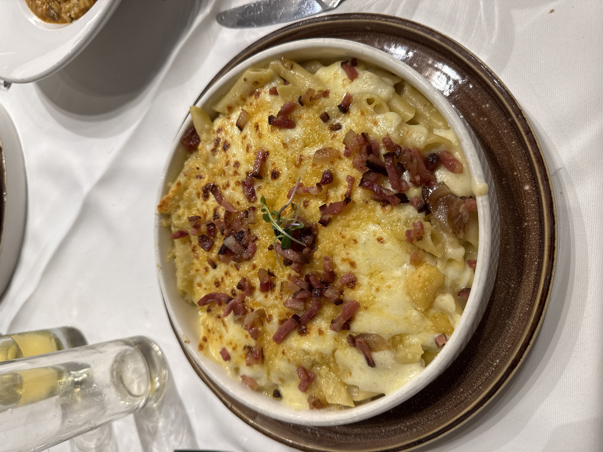 A close-up, overhead shot depicts a bowl of pasta gratin topped with bacon bits, garnished with a microgreen sprig. The pasta is served in a white, circular bowl set on a brown plate. The scene is set on a white tablecloth-covered surface. Other dining elements, including a glass tube and the corner of another dish, are visible in the periphery. The setting appears to be a restaurant. While the information is insufficient to confirm the location, the prompt mentions Caracas, Venezuela.