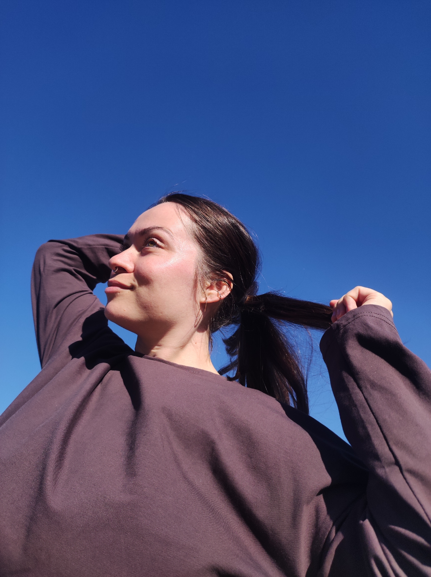 A woman with dark hair pulled into a ponytail, wearing a long-sleeved plum-colored shirt, is outside under a clear blue sky. She is holding her ponytail with her right hand and has her left hand raised to the back of her head. She is looking upwards to the left of the frame. The location is not evident from the visual information.
