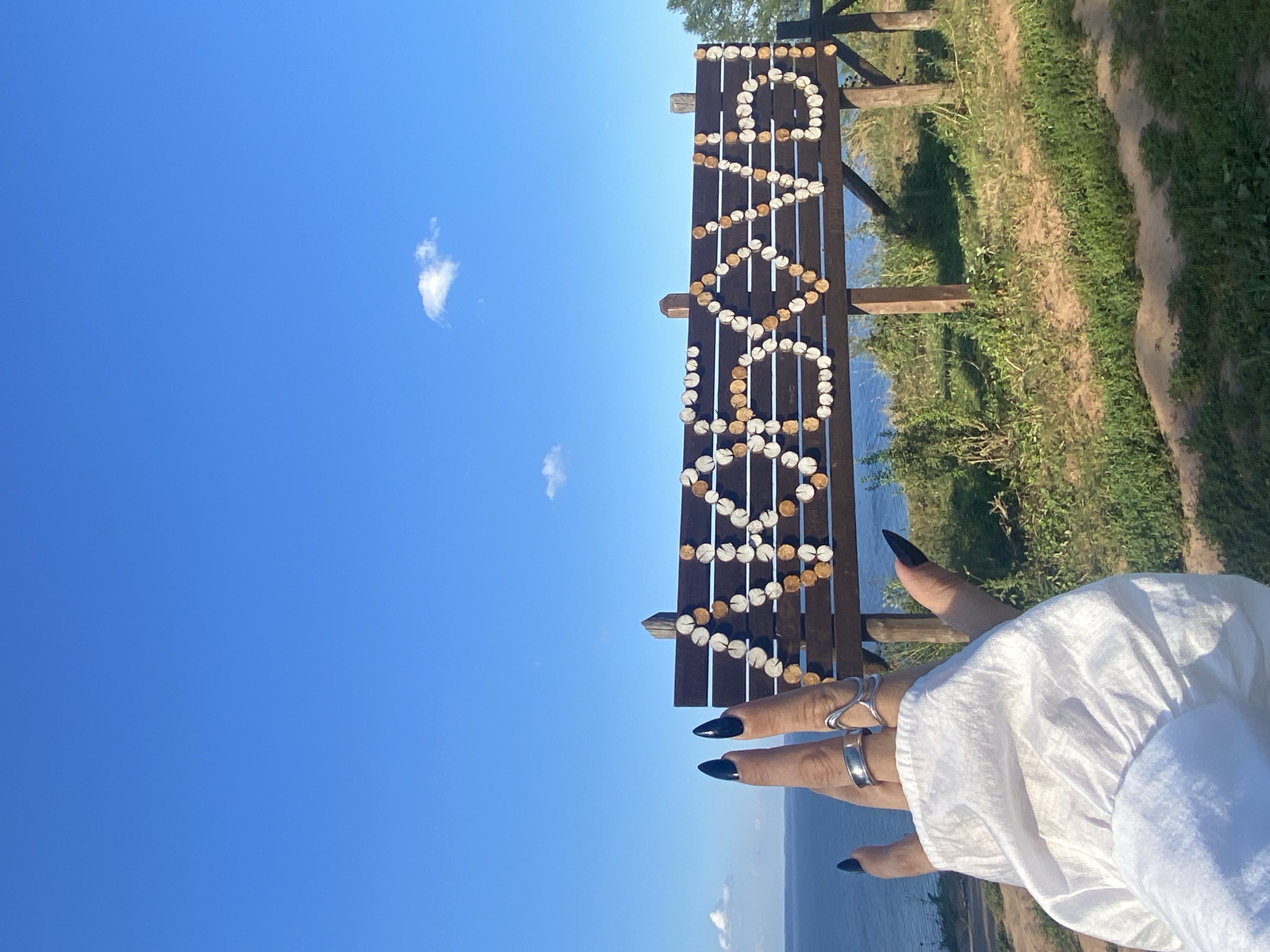 The image shows a vertical shot of a wooden sign adorned with round wooden pieces, forming the word "KOSTOVATY". The sign is placed outdoors, with a clear blue sky and some white clouds in the background. To the right of the sign, there is a glimpse of a body of water and lush green vegetation on a hill. In the foreground, a person's hand with long, black painted nails and several rings is visible, reaching towards the sign. The hand is partially covered by a white, textured garment, likely a shirt or jacket. The lighting suggests it is daytime, possibly late morning or early afternoon, given the brightness of the sky and the shadows. The scene appears to be a scenic overlook or a park, given the presence of the sign and the natural landscape. The overall mood is relaxed and inviting, suggesting a tourist or recreational setting.