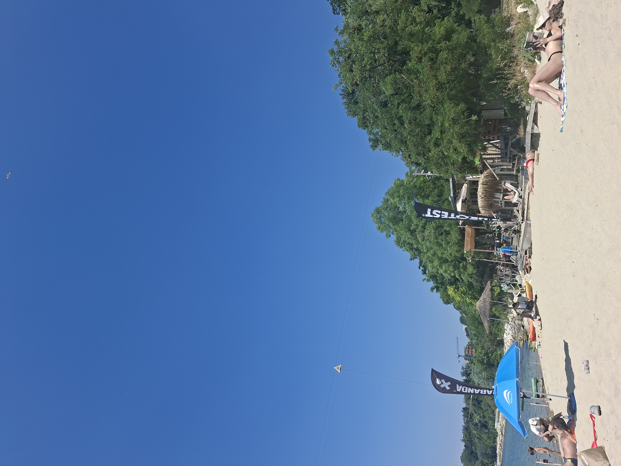 This media file depicts a sunny day at a beach in Varna, Bulgaria. The scene is vibrant and active, with a clear blue sky dominating the upper portion of the frame.
In the foreground, a blue beach umbrella shades a couple of people, one of whom appears to be a child. To the left of the umbrella, a black flag with white text reads "SABANDA X". Further back, a thatched umbrella provides shade to a few individuals, and a small building or structure can be seen nestled amongst lush green trees.
The sandy beach stretches along the coastline, with several kayaks and other beach equipment scattered around. In the middle ground, a person is lying on a towel, reading a book, while another person, wearing a bikini, is also sunbathing. The overall atmosphere is relaxed and summery.
Further into the background, a person is lying on the sand, and some wooden structures are visible. The image also shows a kite in the sky, adding a touch of playful activity to the scene. The presence of a construction crane in the distance suggests ongoing development or activity in the area. The lighting indicates it is likely midday, with bright sunshine casting clear shadows. The overall impression is one of a lively and enjoyable beach day.
