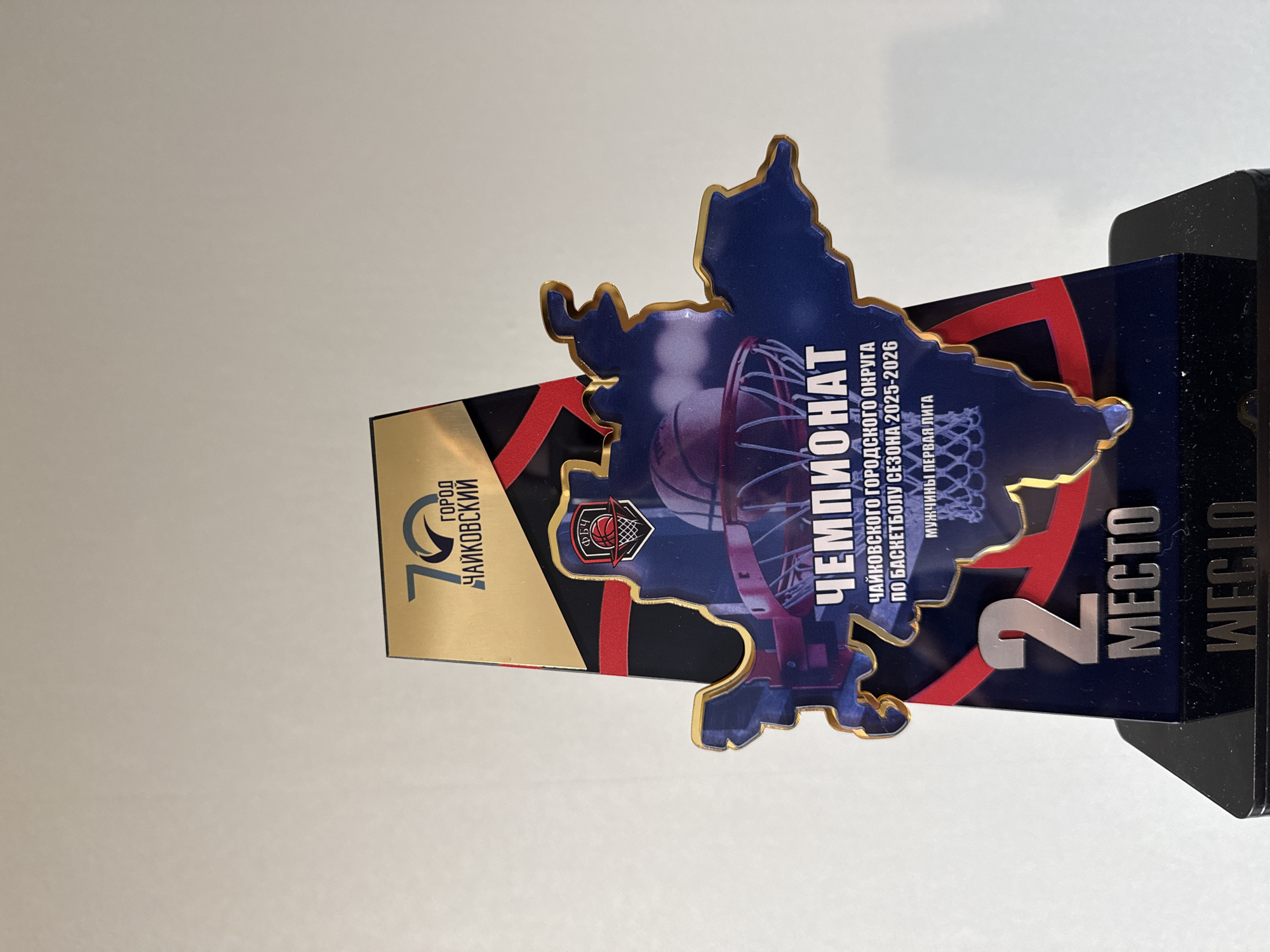 Here is a description of the image:
The image shows a trophy commemorating the "Championship of the Chaikovsky City District" in basketball for the 2025-2026 season, men's first league. The trophy indicates a 2nd place finish. The city of Chaikovsky is located in Russia. The trophy features a map-shaped design element with a basketball net and ball. The object is resting on a light-colored surface.