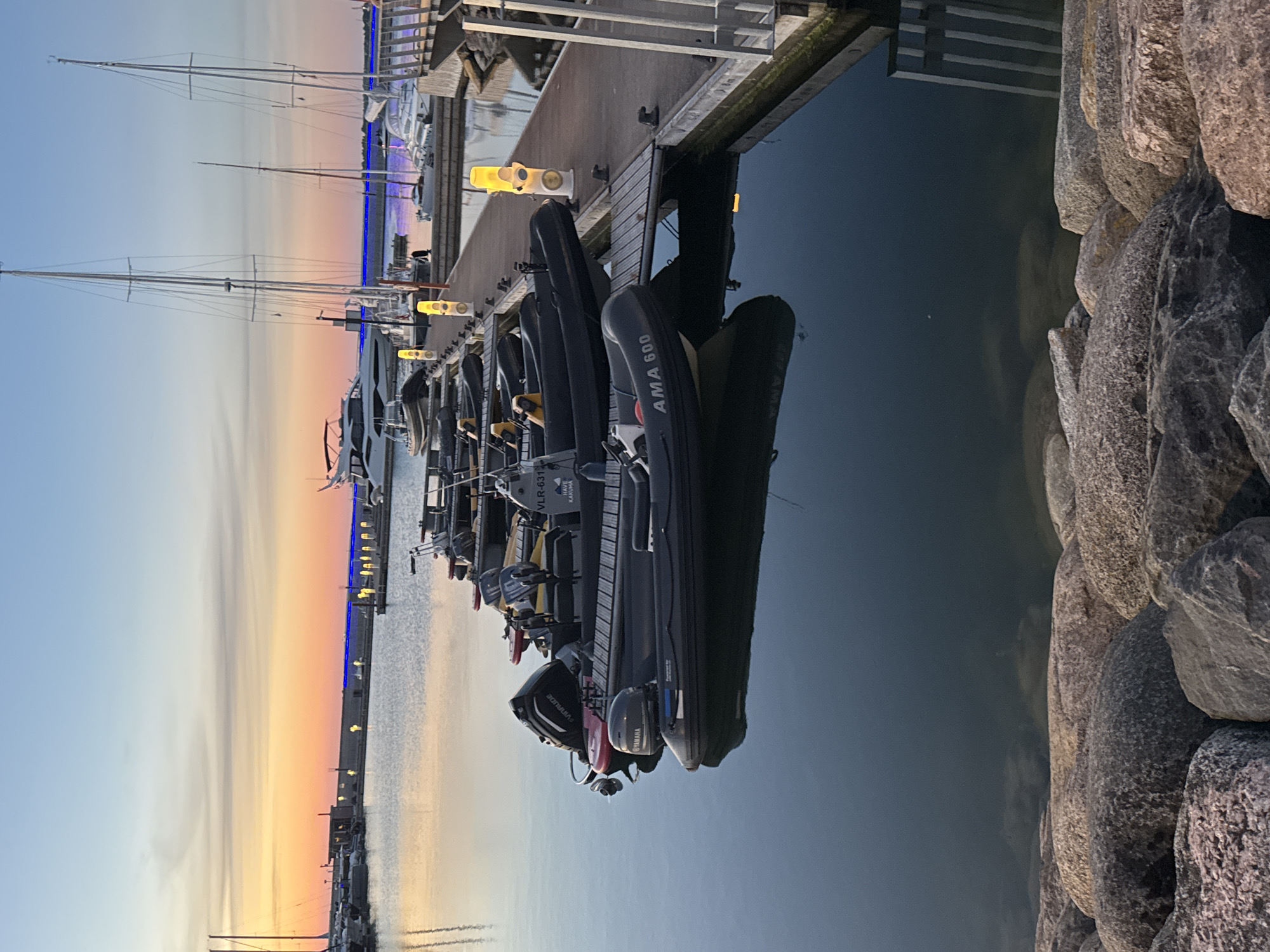 This is a full, eye-level shot of a harbor at dusk, with a sunset visible on the horizon. The image focuses on a row of inflatable boats docked on a wooden pier. The boats are dark gray, some with white lettering. One boat in the foreground is marked "AMA 600". Another boat behind it has "VLR-631" and "HAVE KARUMA" printed on it. There are yellow lights illuminating the pier. In the background, sailboats and a larger white yacht are moored. The water is calm, reflecting the sky and the boats. Large rocks line the foreground, partially submerged in the water. The sky displays hues of orange, pink, and blue, typical of a sunset. The scene appears to be in a marina, likely in Tallinn, Estonia, given the presence of similar harbors and the aesthetic common to the Baltic region. No people are visible in the image.