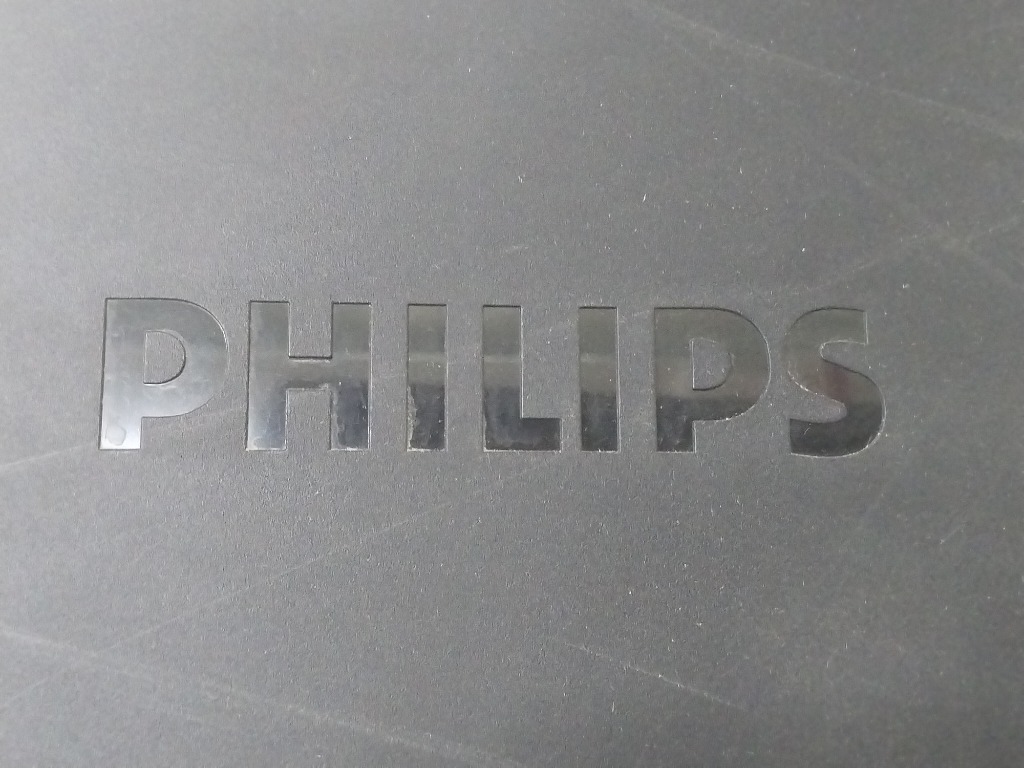 The image is a close-up shot of the word "PHILIPS" embossed on a dark gray, textured surface. The letters are in a metallic silver color and appear to be recessed into the surface. The lighting casts subtle shadows, highlighting the embossed nature of the lettering. The background is uniformly dark gray with some faint scratches and dust specks visible, suggesting the object is likely a piece of electronics or an appliance. There are no people or any other distinguishing environmental features visible in the frame.