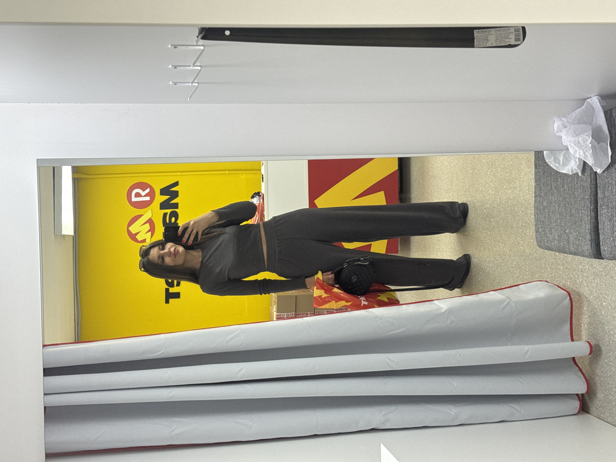 A full-length image of a woman wearing a long-sleeve dark gray top and matching dark gray pants, holding a black purse in her left hand, is reflected in a mirror. The woman is holding a phone in her right hand. The mirror is in a fitting room with a drawn white privacy curtain with a red trim. A wall with the logo "WSM" is visible behind the woman in the reflection. The area beyond the fitting room also contains boxes. The location is Chaikovsky, Russia. A folded paper or tissue is visible near the upper right corner. A black track with hooks for the privacy curtain is visible in the upper center.
