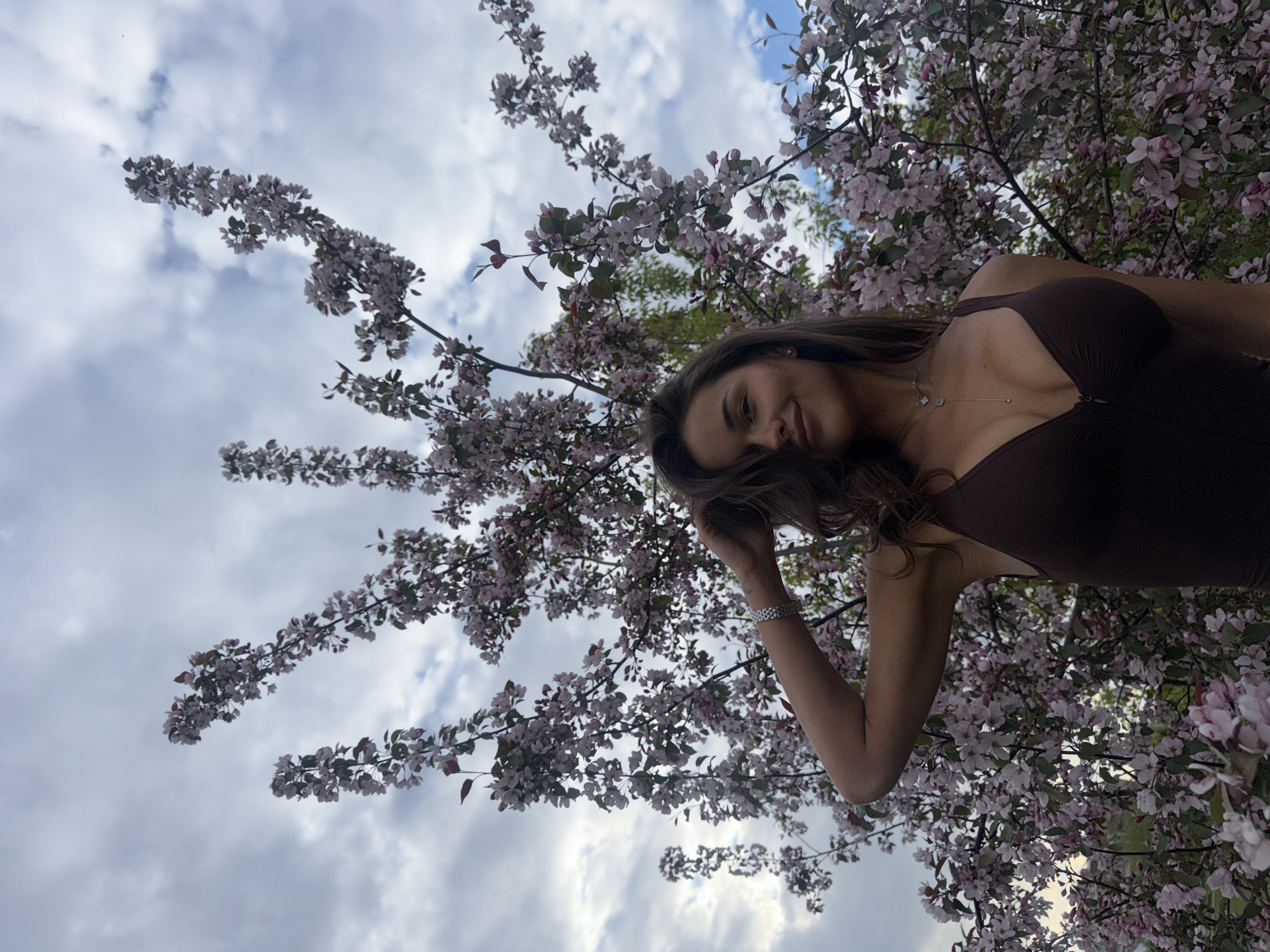 A young woman with long brown hair stands beneath a flowering tree with pink blossoms and green leaves. She wears a brown sleeveless top, a necklace, and a bracelet on her left wrist. Her left hand is near her hair. The background includes a partly cloudy sky.
The location is identifiable as Krasnoyarsk, Russia, due to the flowering trees in this area.