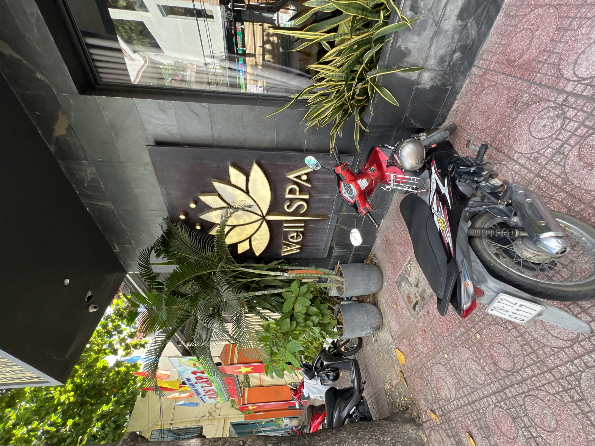 The image shows the exterior of a spa named "Well SPA" located in Nha Trang, Vietnam. The spa has a dark, textured facade with a large sign featuring a golden lotus flower logo and the spa's name in gold lettering. In front of the spa, there are several potted plants, including palm trees and snake plants. Two motorbikes are parked on the sidewalk. The sidewalk is paved with red tiles in a patterned design. In the background, there is a building with colorful flags and signage, suggesting it might be a school or a community center. The scene appears to be a typical urban street in Vietnam, with lush greenery contrasting with the modern spa facade.