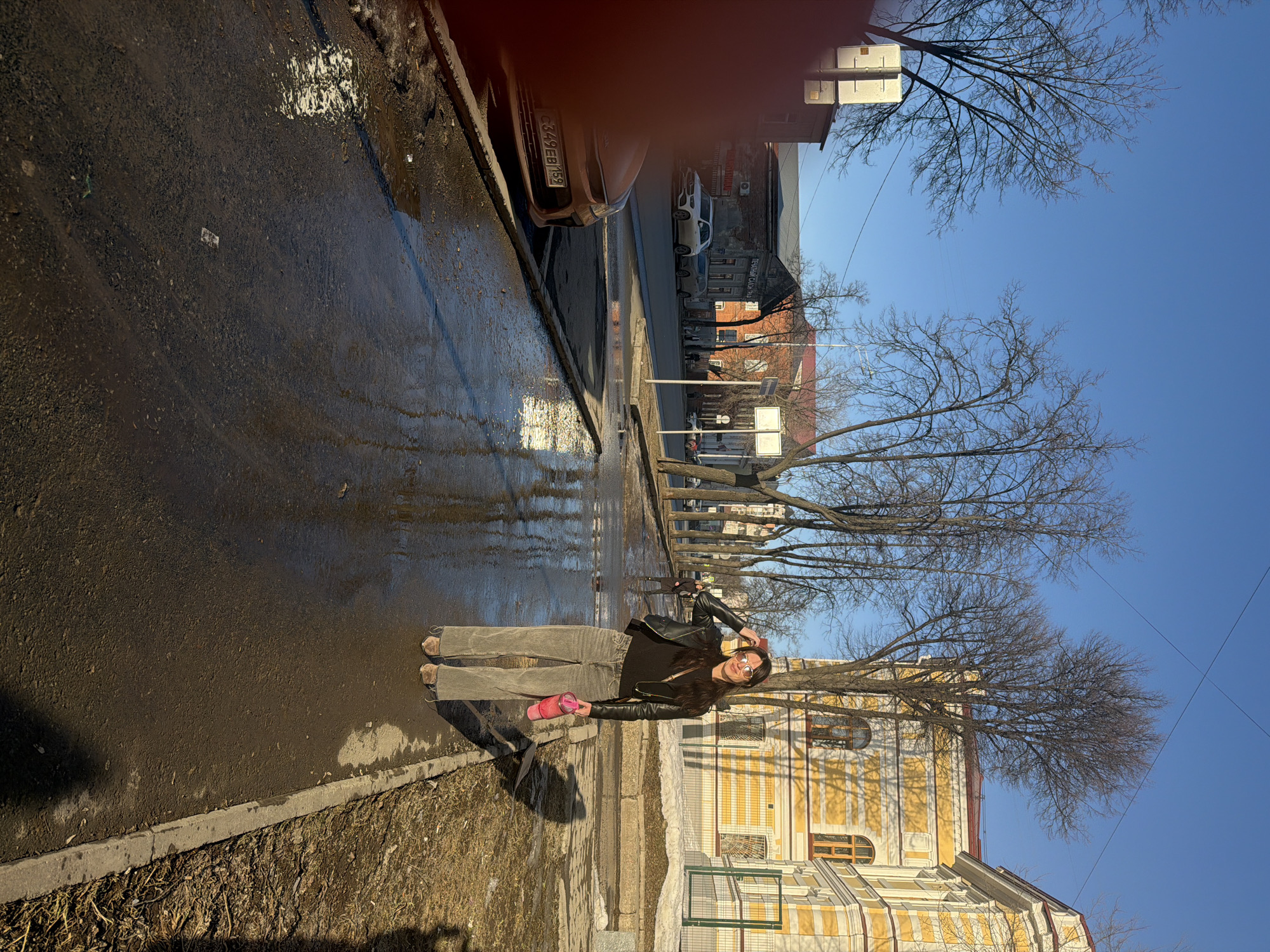 A young woman stands on the sidewalk next to a wet street on a sunny day. She is wearing a black leather jacket, a dark shirt, and corduroy pants, and is holding a pink water bottle. Behind her is a yellow and white building with arched windows. In the background, there are trees without leaves, a car with a license plate beginning with "C349EB159", and buildings lining the street. The location appears to be in the city of Perm, Russia, judging by the car's license plate.