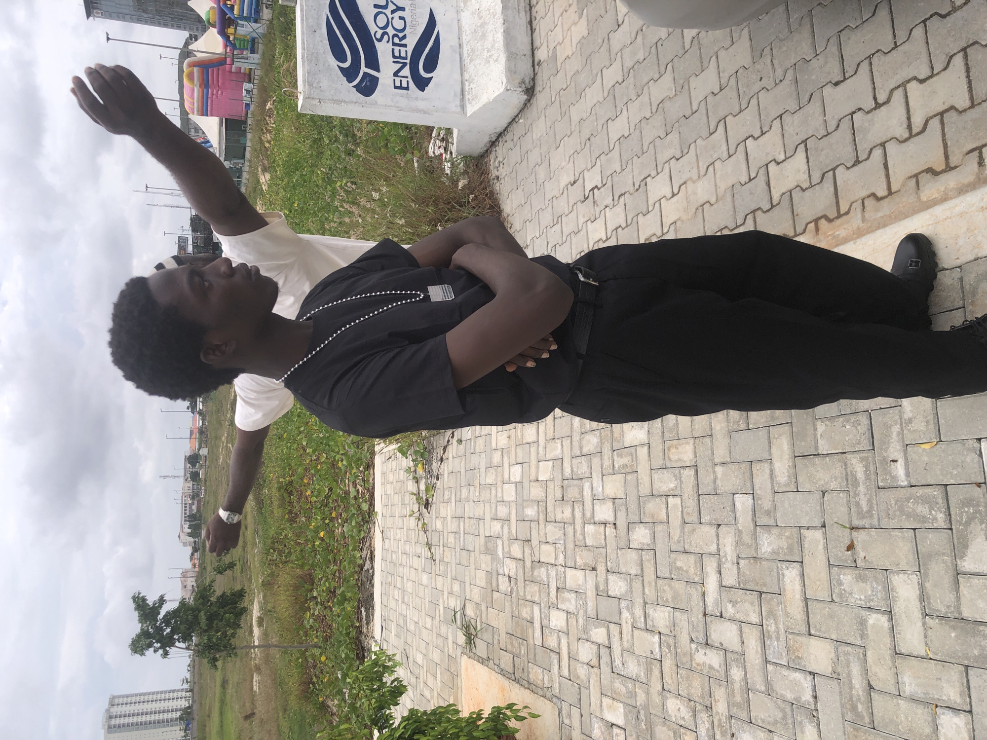 In Ikoyi, Nigeria, a young Black man stands with his arms crossed in the foreground, looking to the right. He wears a black t-shirt, black pants, and black shoes, and a silver chain with a rectangular pendant hangs around his neck. Behind him, another young Black man, mostly obscured by the first, wears a white t-shirt and has his right arm outstretched.
The men are positioned on a paved walkway made of rectangular gray bricks laid in a herringbone pattern. To the right of the men, a concrete barrier with the "SOL Energy Nigeria" logo is visible. In the background, a grassy field with scattered yellow flowers stretches towards a hazy cityscape. Several buildings, including a tall white high-rise and a large inflatable structure with colorful sections, are visible in the distance. The sky is overcast with clouds.