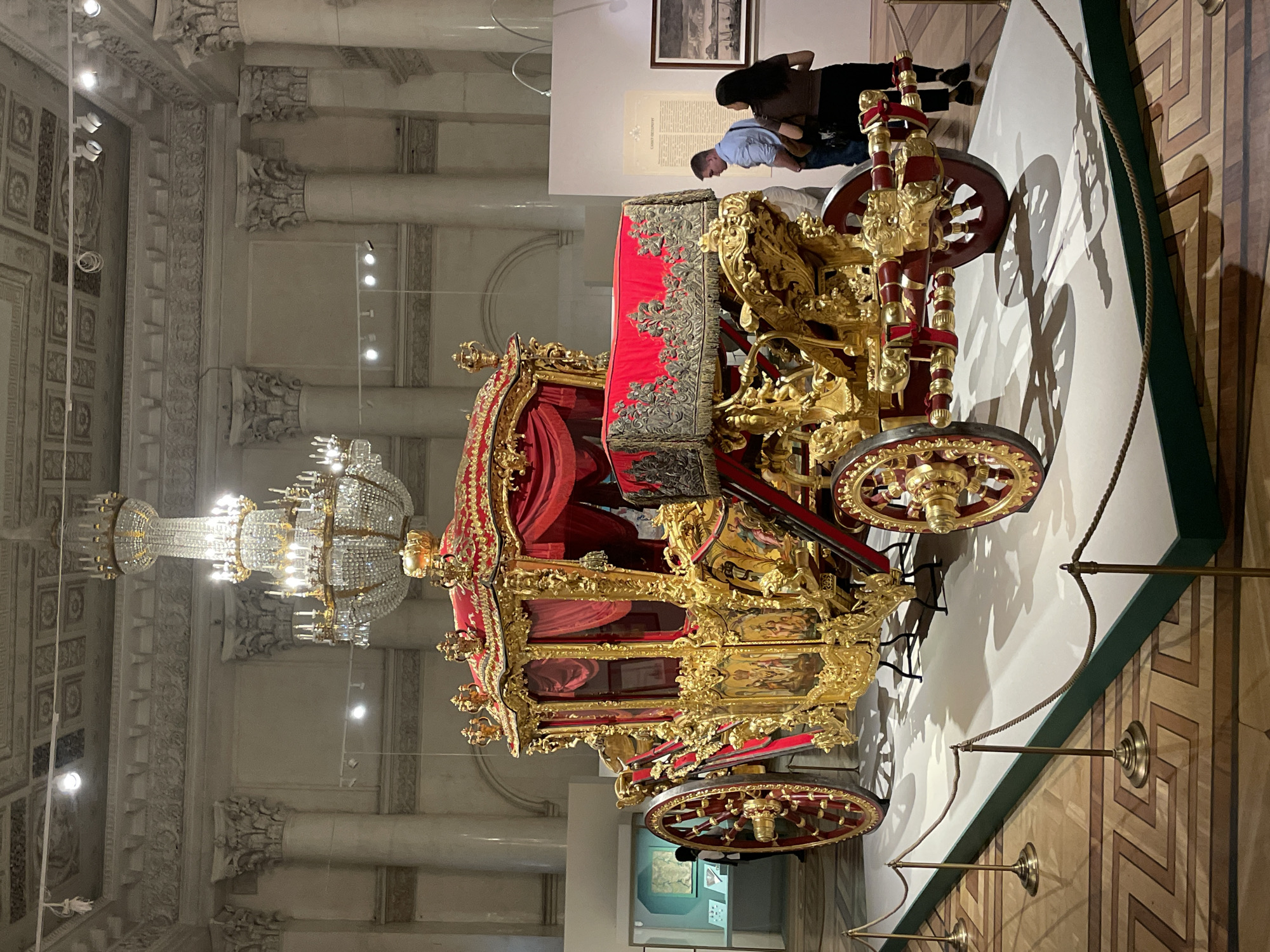 Here's a detailed description of the image:
**Scene:** Interior view of a museum displaying a historic ornate carriage. The scene is well-lit.
**Location:** Unknown city, Unknown.
**Objects:**
* **Carriage:** The dominant object is a highly detailed, gilded carriage with red fabric and intricate carvings.
* **Chandelier:** Large crystal chandelier is visible in the left side of the image.
* **Rope Barrier:** A rope barrier supported by metal stands surrounds the carriage.
* **Display Sign:** A sign with text and images is visible behind the carriage.
* **Wall Art:** In the background above the carriage, there are framed images and documents displayed on the wall.
* **Architecture:** The architecture includes detailed columns and decorative ceiling.
**People:**
* A pair of individuals are standing towards the back. One person in a blue shirt appears to be reading a display, while the other, with long dark hair, is standing next to them.
**Environment:**
* The floor has a geometric pattern.
* The room has high ceilings.
* The walls are light colored, providing contrast for the displays.
**Action/Interaction:**
* The pair of people are observing the displays.
* The museum is set up to prevent close access to objects with rope barrier.