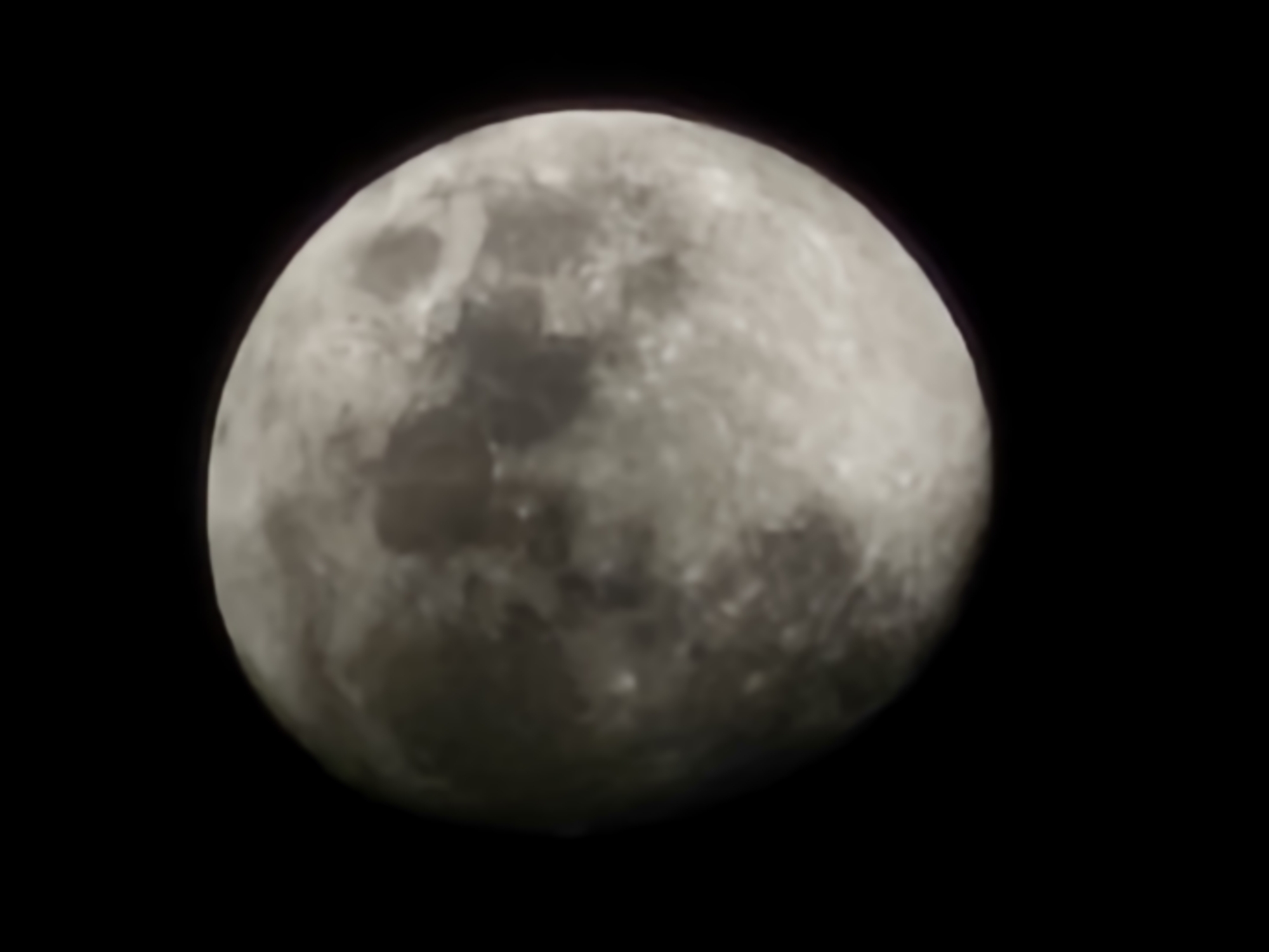 An astronomical photograph captures the full or near-full Moon against a stark, black background. The Moon is the sole subject, presented as a spherical, three-dimensional object dominating the frame. Its surface is clearly visible, showcasing a varied topography of lighter, heavily cratered highlands and darker, smoother maria (lunar seas).
The image emphasizes the Moon's mottled appearance with distinct grey tones, suggesting differences in composition or light reflection across its surface. Notable details include the slightly soft focus or atmospheric blurring, which lends a somewhat ethereal quality to the celestial body. Along some edges of the Moon, particularly where it meets the dark void of space, a subtle reddish-purple fringing, indicative of chromatic aberration, can be observed. The completely black background suggests either a deep night sky free of light pollution or the vacuum of space, highlighting the Moon in isolation. There is no visible activity or event taking place, as it is a still photographic capture of a celestial body. No text is visible in the image.