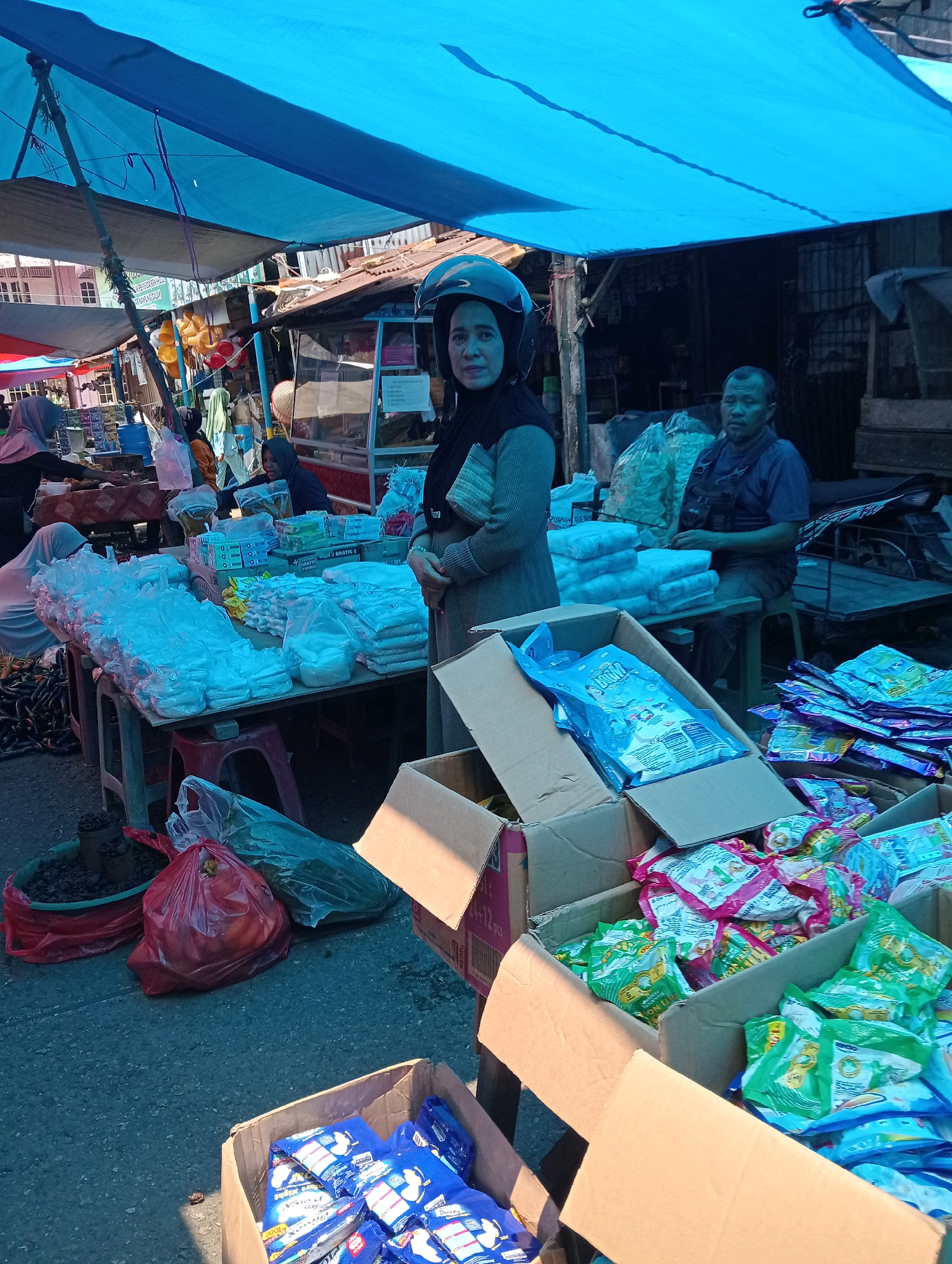 This image depicts a street market scene in Pariaman, Indonesia. A woman wearing a black hijab and a motorcycle helmet stands in the foreground, looking towards the camera. She is wearing a grey knitted sweater. Behind her, a man in a blue shirt and dark overalls sits on a stool. The market is covered by a large blue tarp. Tables are laden with various goods, including numerous plastic-wrapped packages, some in boxes. Bags of produce are also visible, including a red plastic bag containing what appear to be fruits and a green bag filled with dried items. The ground is asphalt. The overall impression is of a bustling, everyday market.