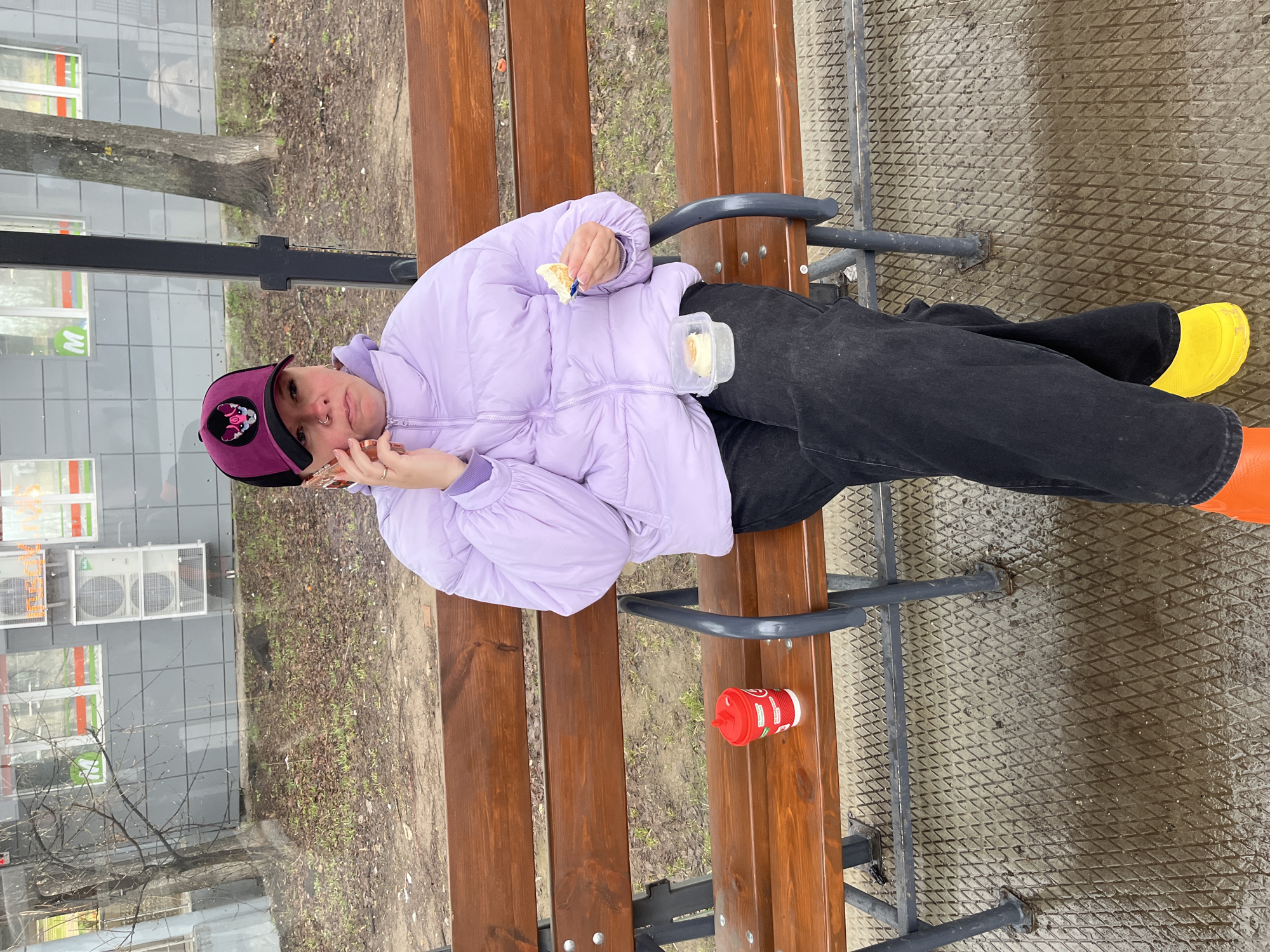 A young individual is captured sitting sideways on a sturdy wooden and metal public bench in an outdoor setting. They are dressed in a light purple puffer jacket, black pants, and a dark purple baseball cap featuring a distinctive emblem, likely a cartoon character. A nose ring is visible on their face. The person is engaged in two activities simultaneously: holding a small piece of light-colored food to their mouth with one hand while holding a phone to their ear with the other, suggesting they are eating a snack and talking on the phone. Their feet, adorned with bright yellow and orange boots, are visible resting on the ground.
On the bench beside them, a small, clear plastic container holds additional portions of the food. A red disposable cup with a white lid and straw is also placed on the bench. The ground directly beneath the bench features a dark, textured metal grate, which appears damp. Beyond this, the ground transitions to dirt, scattered dry leaves, and patches of new green growth, suggesting a cool or damp season, possibly late winter or early spring. A bare tree stands in the mid-ground. In the background, a modern building with gray tiled walls is visible, featuring several external air conditioning units. Windows on the building display a prominent "M" logo, possibly indicating a grocery store or commercial establishment. The time of day is daytime, under an overcast sky. The overall atmosphere is casual and everyday, set in what appears to be an urban or suburban public space in Chaikovsky, Russia. The text on the red cup is not clearly legible.