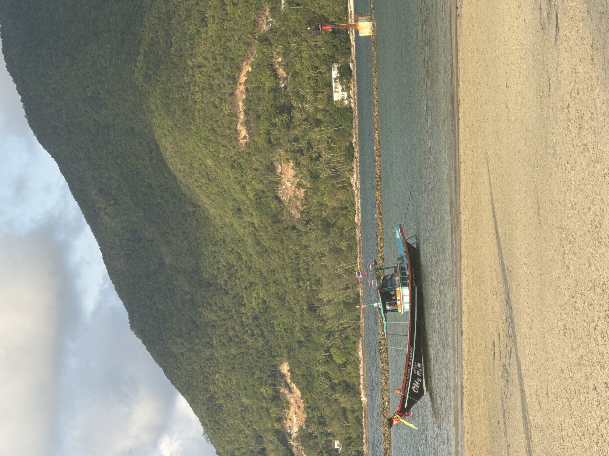 A ground-level, outdoor photograph captures a coastal scene in Baan Chaloklum, Thailand, featuring a traditional Thai longtail boat at rest near the shore. The boat, characterized by its dark hull and brightly colored accents, is positioned parallel to a sandy beach. The boat has Thai flags and other decorations, and writing along the hull.
The backdrop comprises a steep, densely forested mountain, creating a contrast with the sandy shoreline and the body of water separating it from the boat. A pier structure extends into the water, and what appears to be a breakwater, made of concrete blocks, runs along the shoreline. The sky, seen at the top left, is overcast with grey clouds.
