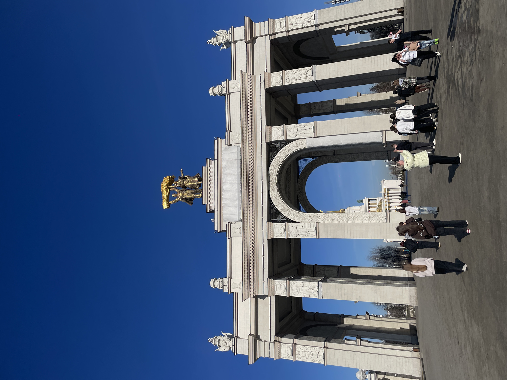 The image shows a monumental archway under a clear, bright blue sky. The archway is constructed of light-colored stone and features detailed relief carvings. Atop the archway stands a golden statue of figures holding a sheaf of wheat. Several people are seen walking through or standing near the archway. The location is the Main Entrance Arch of VDNKh, Moscow, Russia, a landmark known for its Stalinist architecture.