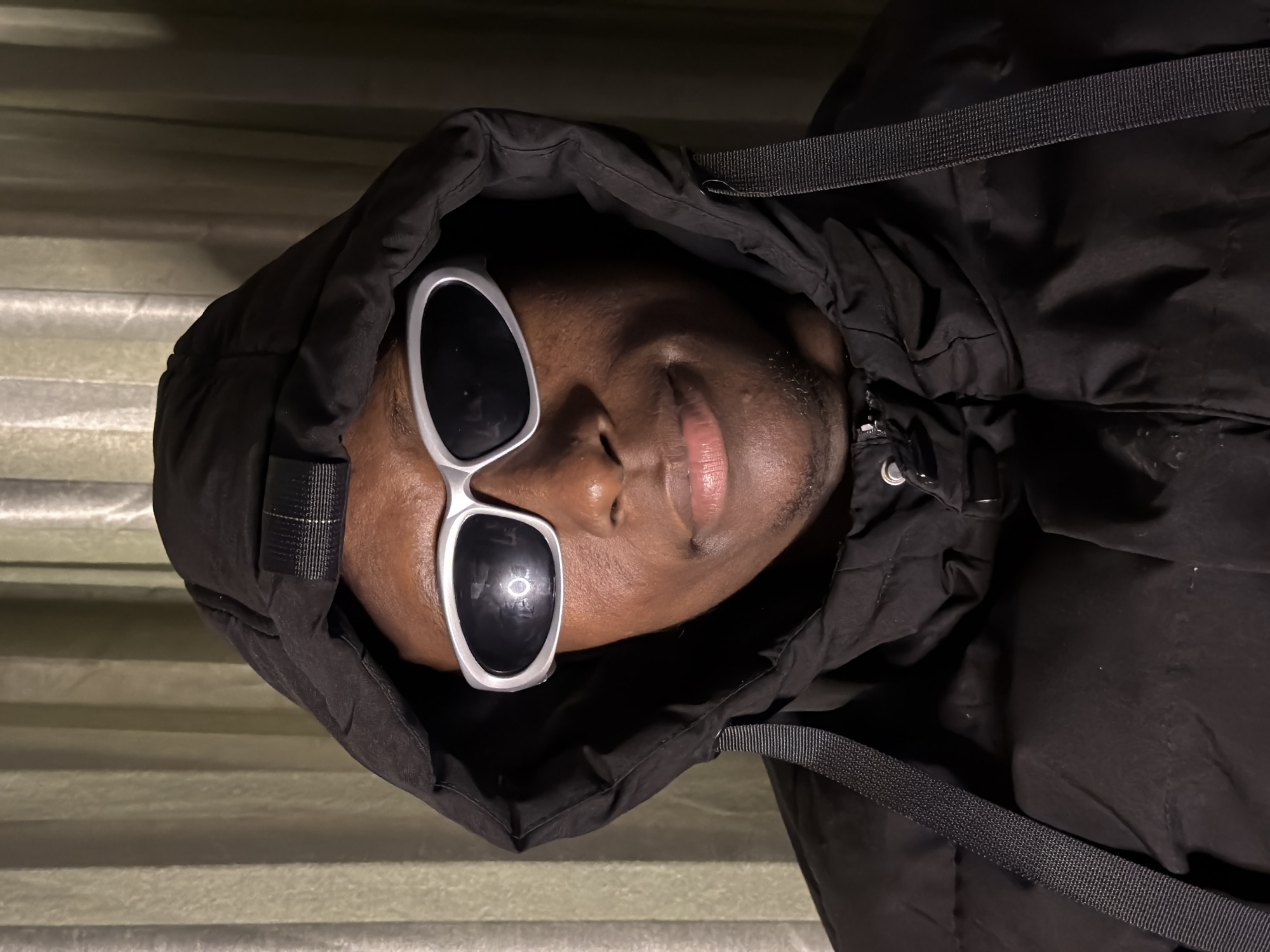 A close-up, medium shot captures a Black individual wearing a black hooded puffer jacket and silver-rimmed, wraparound sunglasses. The person's face is turned slightly to the right, with their lips parted in a slight smile. The background consists of a softly lit, olive-green curtain. The scene is lit from the front, creating a subtle reflection on the lenses of the sunglasses. The image appears to be a self-portrait or a posed photograph. There are no discernible elements in the image that would suggest a specific location within the city of Jiangjin, China.