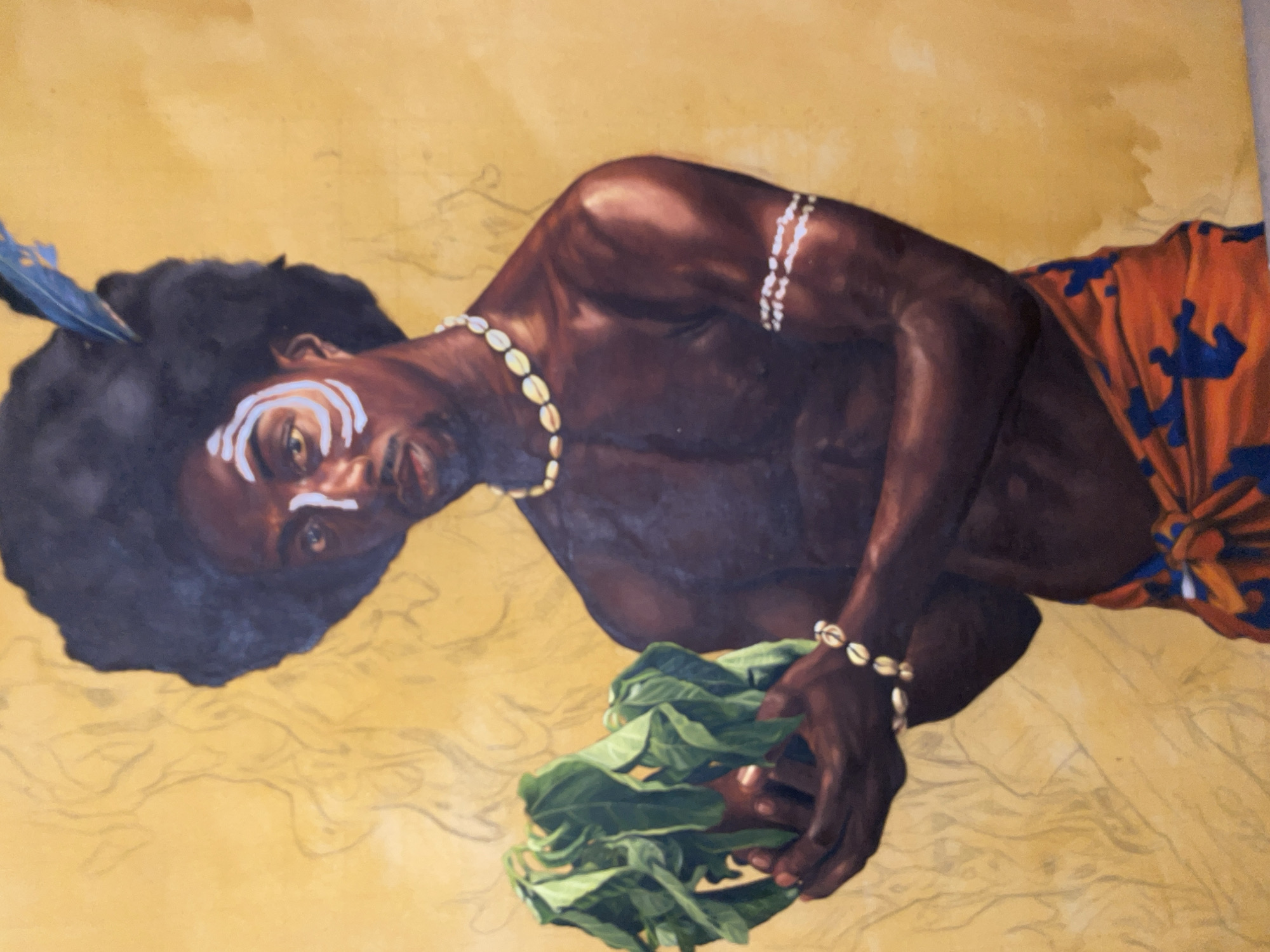 An oil painting depicts a dark-skinned individual, likely male, lying diagonally across a light yellow-brown background. The figure has dark, curly hair adorned with a blue feather on the left side. White ceremonial paint marks their face: three stripes across the forehead and two under the right eye. They wear a necklace made of cowrie shells and a matching bracelet on their left wrist. In their right hand, they hold a bundle of fresh green leaves. An orange fabric with distinct blue patterns is draped around their lower body. The background is largely unpainted, revealing faint pencil sketches that outline additional forms, suggesting the artwork is either unfinished or stylistically incorporates these preliminary lines. The person gazes directly at the viewer with a calm, engaging expression. The artistic style features rich, realistic skin tones and detailed adornments. No text is visible in the image. Given the cultural adornments, the painting likely represents a figure from an African cultural context, consistent with the provided location of Ado, Nigeria.