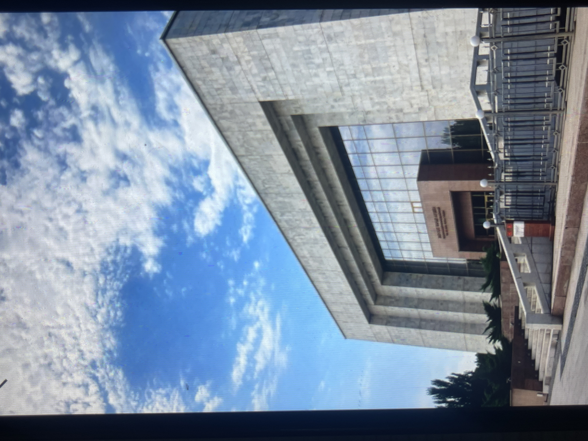 The image depicts the National Library of the Kyrgyz Republic in Bishkek, Kyrgyzstan. The building is a large, modern structure with a facade clad in light-colored stone or marble. A prominent feature is the expansive glass-fronted section, which reflects the bright blue sky dotted with white clouds. In front of the library, there is a set of concrete stairs leading up to the entrance. A dark metal fence with decorative elements and globes tops the fence encloses a portion of the building's exterior. Several trees and bushes are visible in the surrounding area, adding greenery to the scene. The overall impression is one of a grand and significant public building.