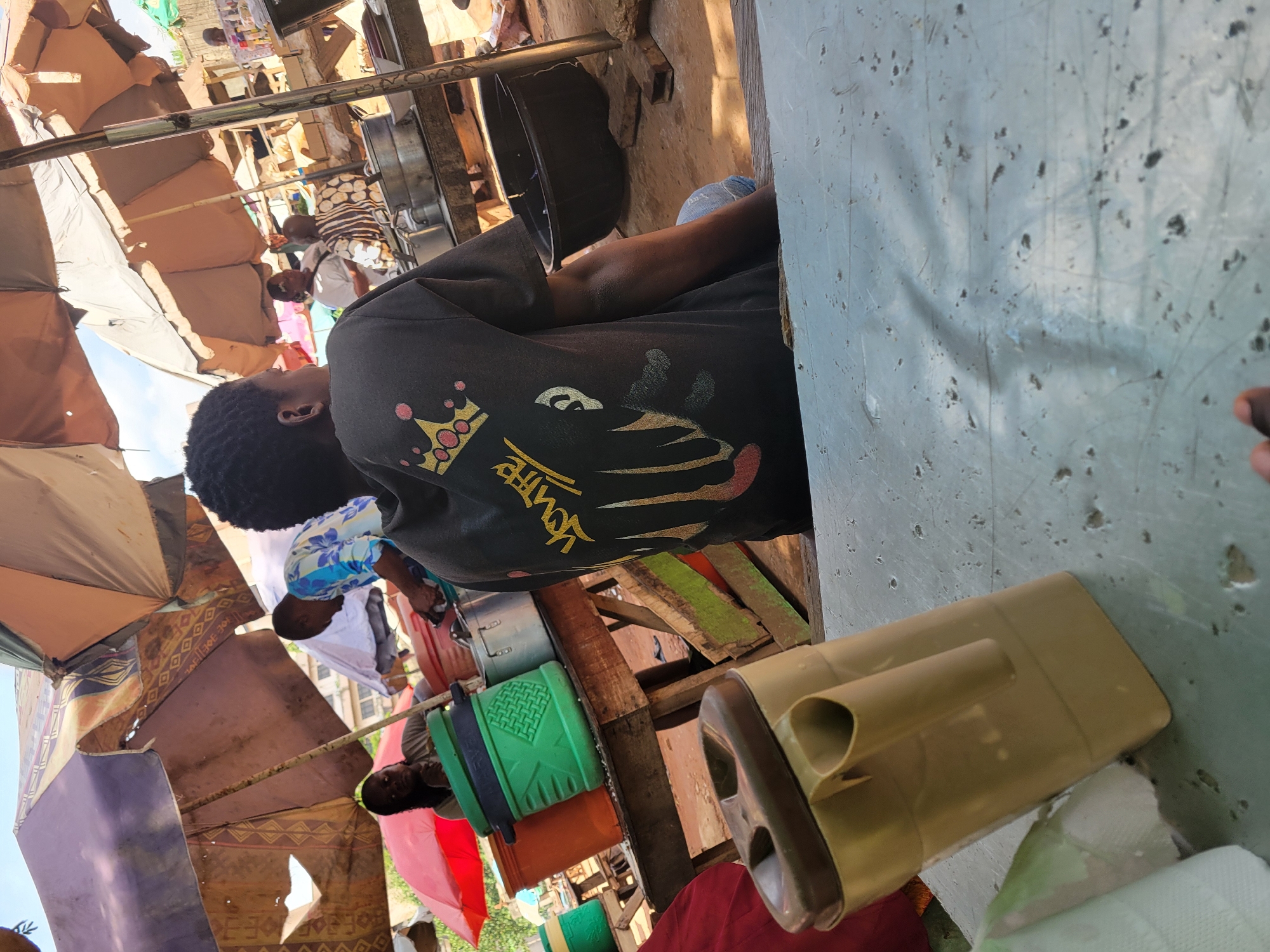This image depicts a bustling outdoor market scene, likely in Abuja, Nigeria, during daylight hours. In the foreground, viewed from behind, a person with short, dark, curly hair is seated at a light-colored, scuffed table. They are wearing a black t-shirt adorned with a prominent golden crown graphic with pink accents, and stylized yellow text that appears to read "YOUTE" or "YOUTH," alongside other graphic elements that resemble abstract shapes or hands.
On the table next to the seated individual is a beige plastic tool with a handle, possibly a scoop or trowel, resting near some crumpled fabric. The table surface shows significant wear and tear.
The background reveals a vibrant and informal market environment. Numerous open-air stalls are visible, constructed from various materials including wood, metal, and what seems to be recycled cardboard or fabric. Large, colorful umbrellas and canopies, some patterned, are spread overhead, providing shade from the bright sun. Several other individuals are present in the market; some are standing or seated in the background, including a person in a white shirt and another in a blue and white floral patterned shirt. Various market items and containers, such as a green plastic bucket and a metallic pot, are scattered around the stalls. The overall atmosphere suggests a busy, community-oriented marketplace.