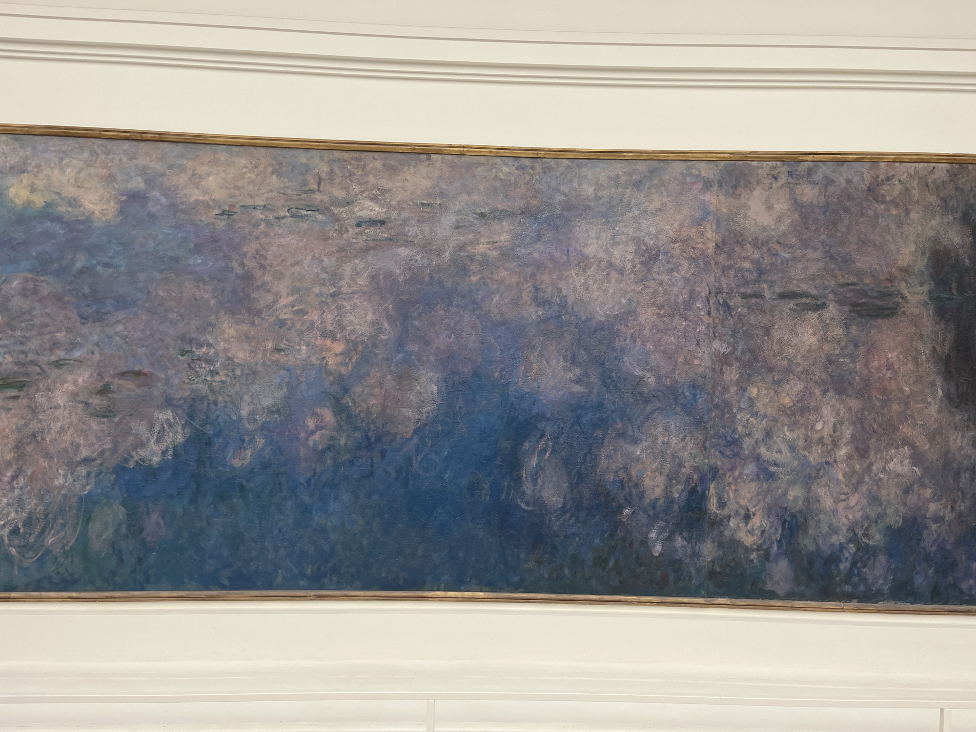 A large, horizontal painting, framed in gold, is displayed prominently on a white wall, indicative of an art gallery or museum setting. The artwork itself is an impressionistic or abstract piece, primarily composed of deep blues, purples, and grays, with swirling, lighter brushstrokes and indistinct forms that strongly suggest a water scene with lily pads, typical of Claude Monet's "Water Lilies" series. The painting's serene, almost ethereal quality is enhanced by its muted color palette. Above and below the framed artwork, elegant white architectural molding adorns the wall. The image captures a static exhibition view, with no human activity or visible text. The presentation style and the subject matter strongly suggest a location such as the Musée de l'Orangerie in Paris, France, known for its immersive display of Monet's monumental water lily panels.