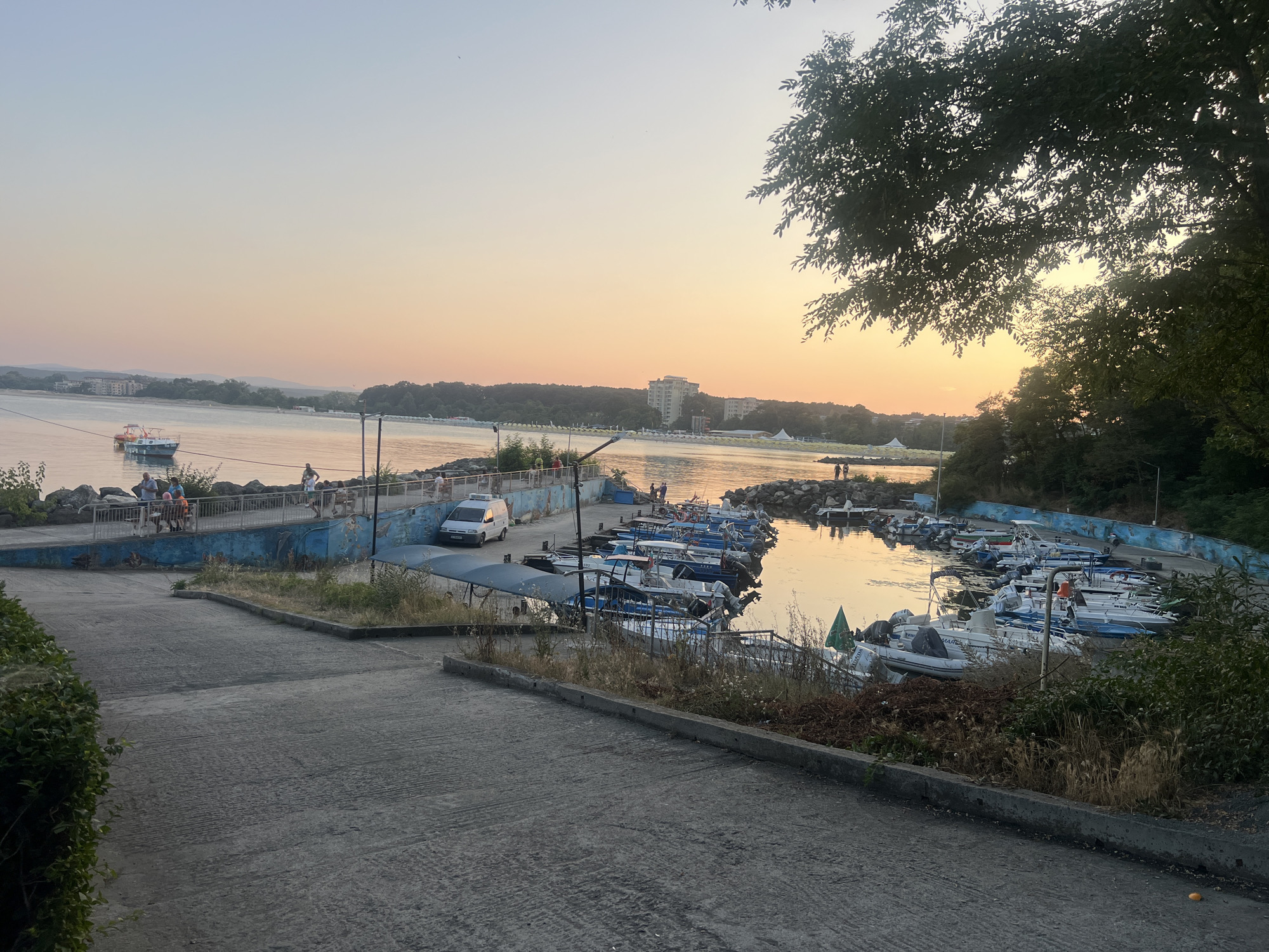 The image depicts a scenic view of a harbor in Primorsko, Bulgaria, during what appears to be sunset. The sky exhibits a gradient of pale blue to warm orange hues, indicative of the setting sun. In the foreground, a paved ramp slopes downwards towards a rocky shoreline. Adjacent to the shoreline is a harbor filled with numerous small fishing boats docked in rows. Some of these boats are white with blue trim, while others are a mix of white and dark colors. A few people are visible on the pier above the harbor, observing the boats or the water.
Further back, across the water, a long stretch of coastline is lined with buildings and lush green trees, suggesting a developed area or town. The water itself is calm and reflects the warm colors of the sky. To the right of the harbor, dense green foliage and trees overhang the scene, framing the view. A white van is parked on the pier near the boats. The overall atmosphere is serene and peaceful, characteristic of a coastal town at dusk.