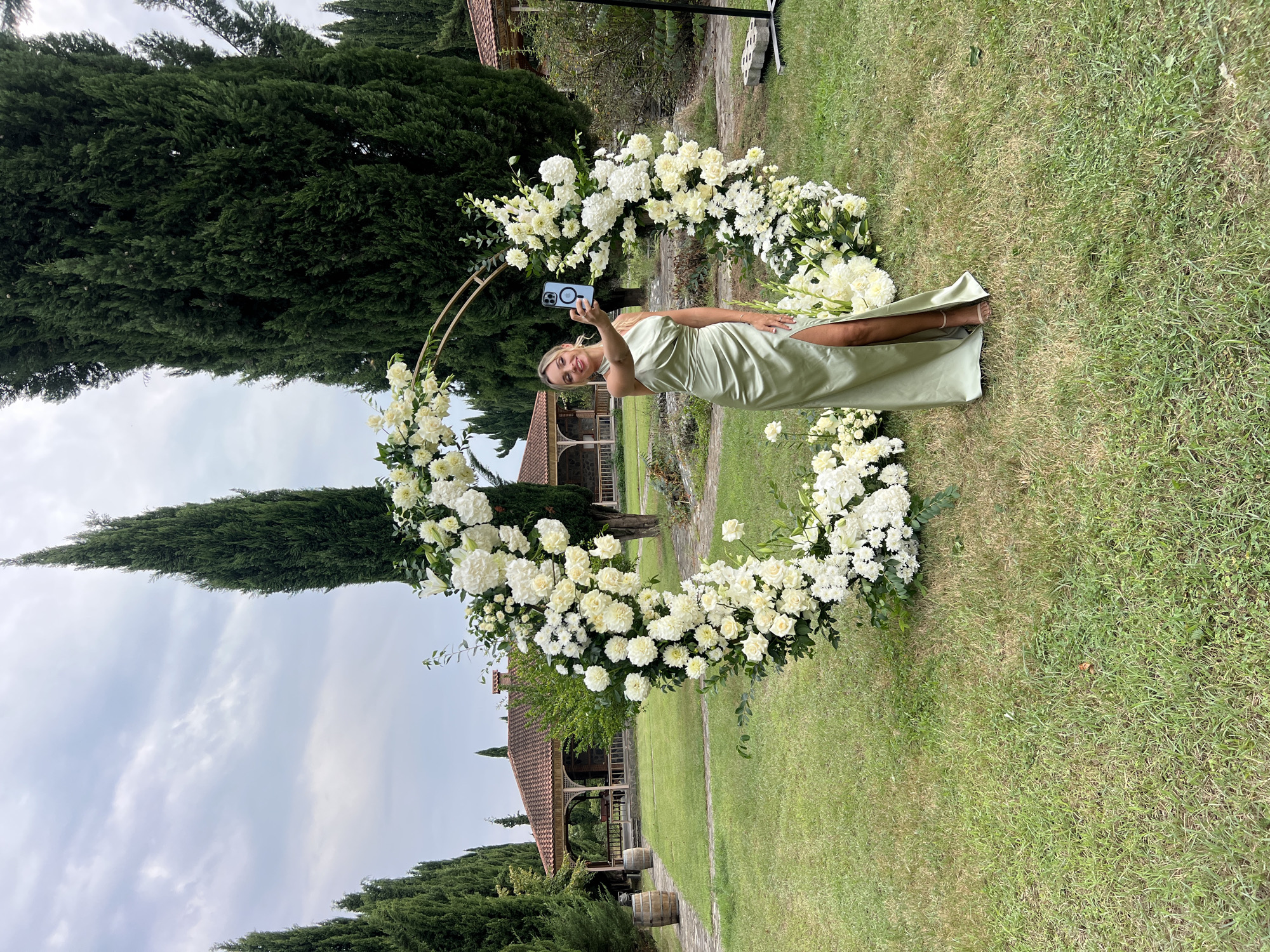 A woman in a green satin dress stands in front of a circular floral arrangement of white flowers. She is holding a smartphone in her right hand, taking a selfie. The scene is set outdoors in a grassy area with tall cypress trees in the background. Behind the trees, there is a building with a tiled roof and open-air verandas, which appears to be part of a resort or winery. Two wooden barrels are visible on the left side of the image, and in the background on the right, there is a black metal stand. The sky is overcast. The location is Saniore, Georgia.