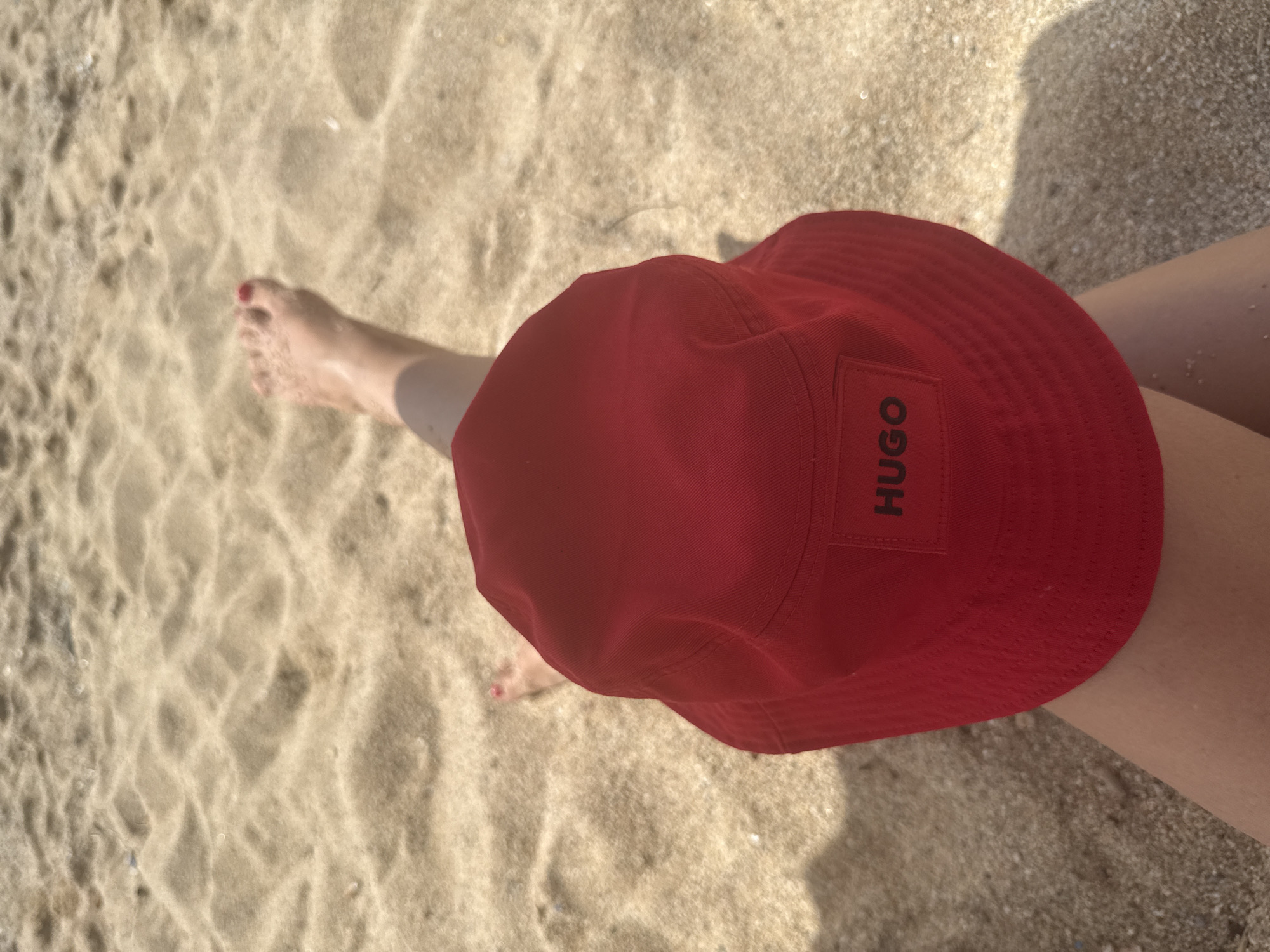 A close-up, top-down shot shows a person's legs and a red bucket hat on a sandy beach. The person is sitting with their legs crossed, and their bare feet are visible in the sand. The hat has a black tag with the word "HUGO" embroidered on it. The sand is light brown and textured, with shadows cast by an unseen object. The scene appears to be a sunny day at the beach. The location is stated to be Badalona, Spain, though no specific landmarks are visible to confirm this.