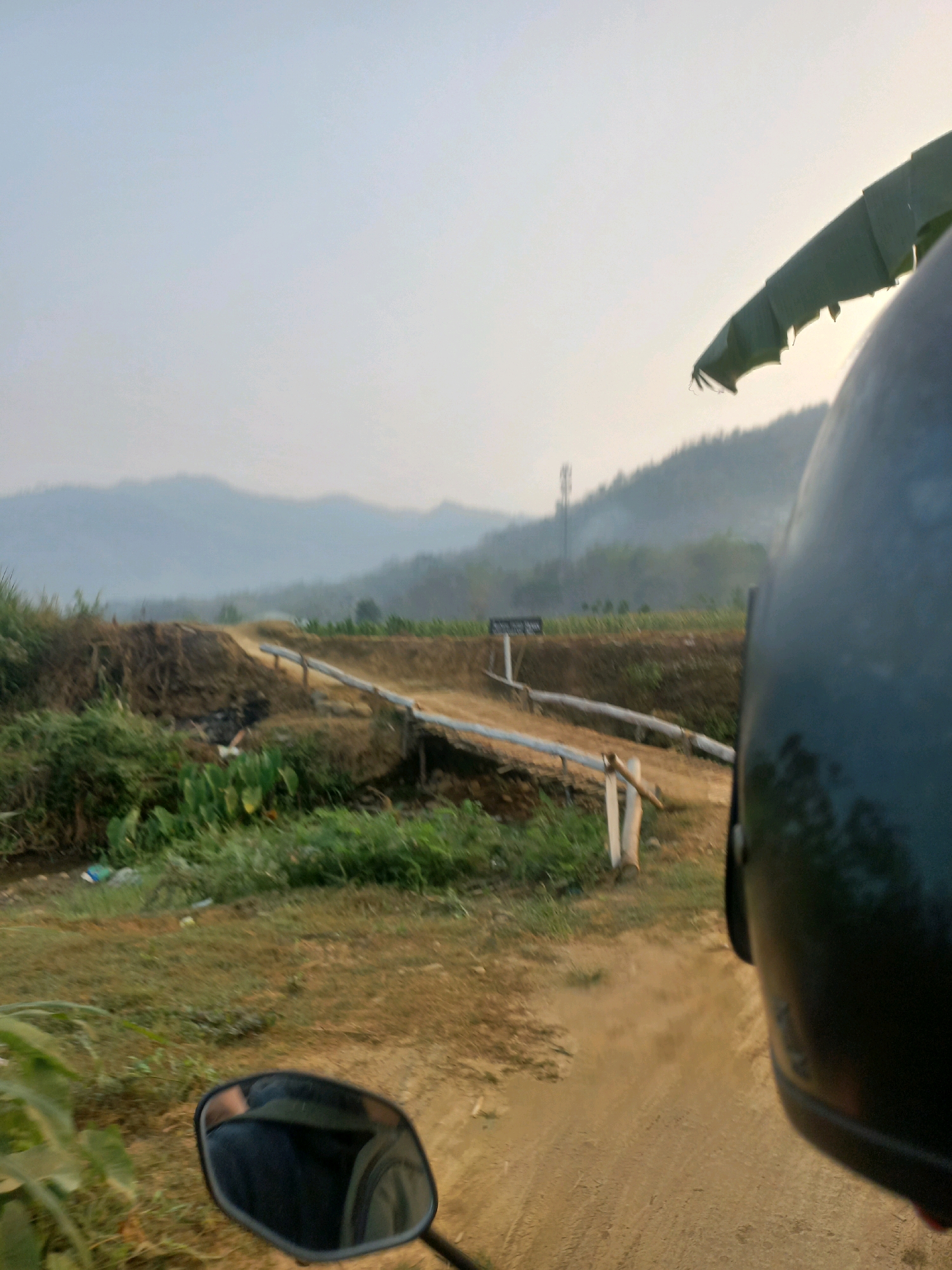 The image depicts a dusty, unpaved road winding through a rural landscape, likely on the outskirts of Wiroko, Indonesia. In the foreground, a black motorcycle helmet dominates the right side of the frame, obscuring the rider's perspective. A rearview mirror, reflecting a dark helmet and possibly the rider's back, is attached to the motorcycle and visible in the lower left quadrant. The road appears to be under construction or undergoing maintenance, with piles of dirt and debris along its edges. A makeshift barrier, consisting of wooden posts and a horizontal pipe, crosses the road, suggesting a closure or a point of controlled passage.
Green vegetation, including tall grasses and broader-leafed plants, lines the left side of the road. Beyond the immediate roadside, a hilly terrain stretches into the distance, shrouded in a light haze, possibly due to atmospheric conditions or agricultural burning. Bare trees with a conical shape dot the slopes of the hills, and a tall communication tower is visible in the middle ground. The sky is overcast, with a soft, diffused light that suggests either early morning or late afternoon. The overall impression is one of a rural, developing area.