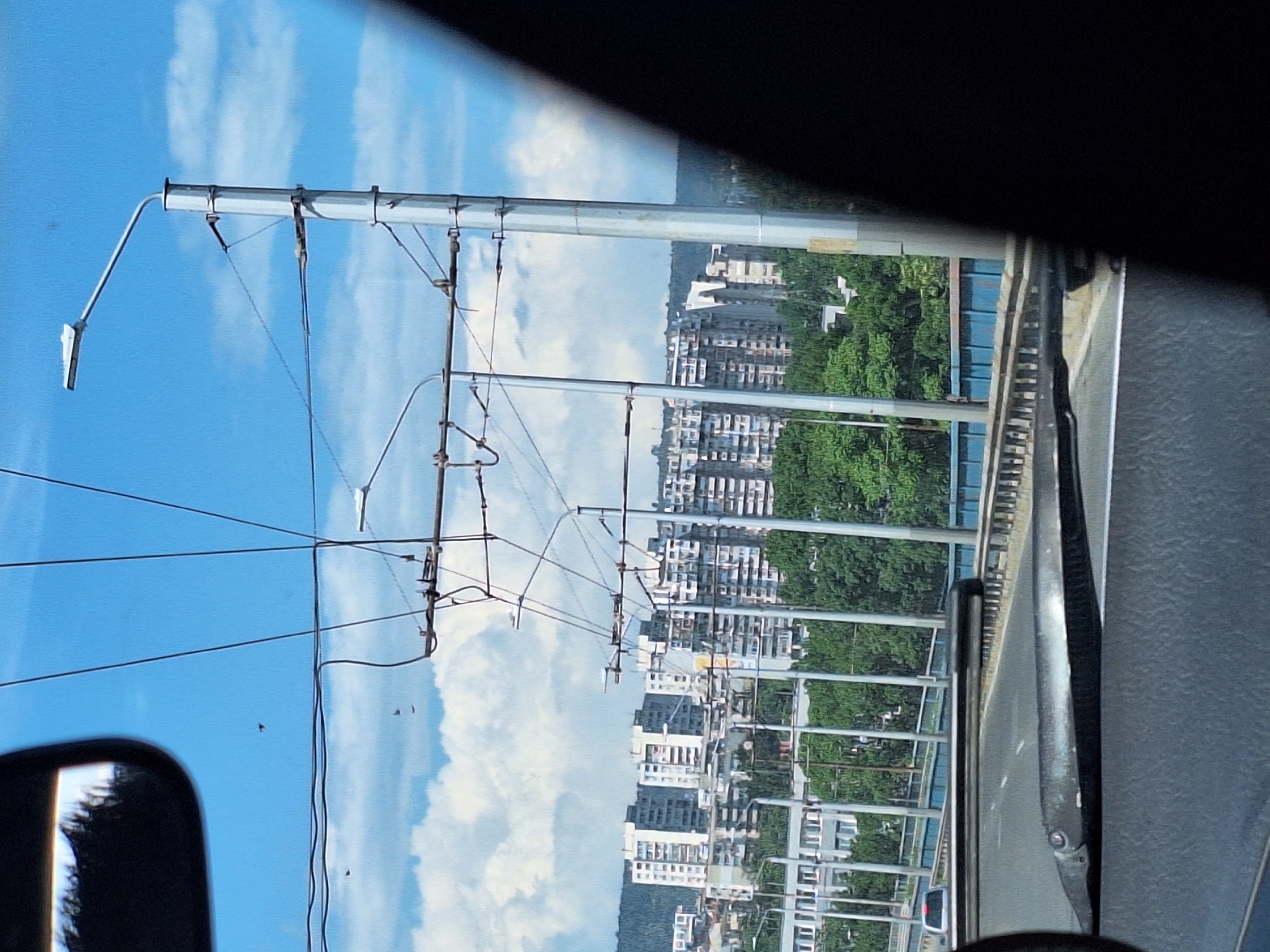 The image captures a view from inside a vehicle on a bridge, likely a highway overpass, in Varna, Bulgaria. The foreground shows a car's windshield wipers and the road ahead, blurred by motion. In the middle ground, there is a blue barrier along the side of the bridge, with trees and poles visible behind it. The background reveals a cityscape with numerous tall apartment buildings, some of which appear to be unfinished or in a state of disrepair. The sky is blue with scattered white clouds. Overhead, a complex network of wires, likely for trolleybuses or trams, is supported by poles. Several dark, small shapes, possibly birds or insects, are visible in the sky.