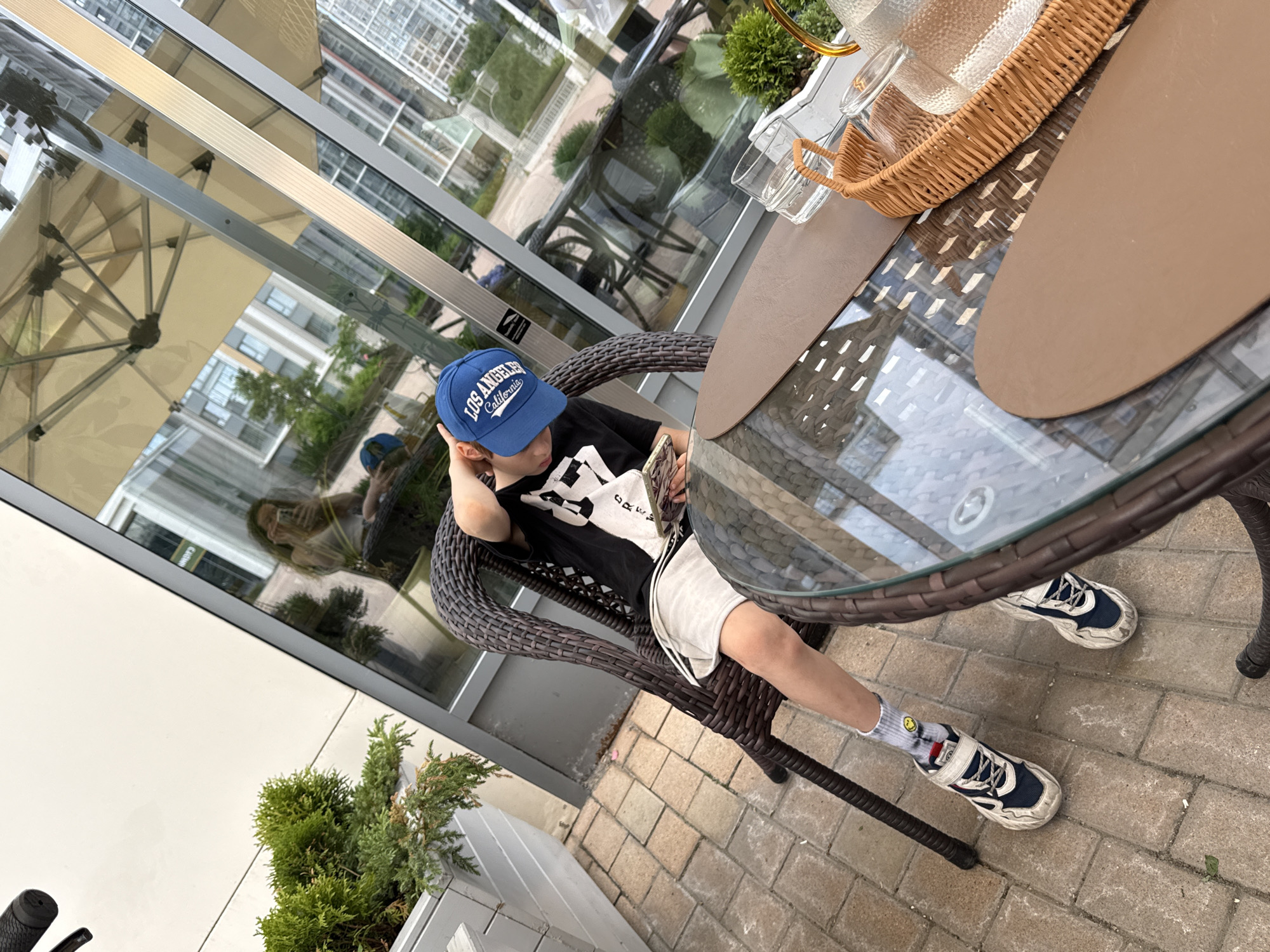A young boy is seated at an outdoor table in what appears to be a patio or cafe setting in Moscow, Russia. He is the central subject, positioned slightly off-center to the right, looking down at a mobile device held in his hands. He wears a blue baseball cap with "LOS ANGELES California" in white text, a black t-shirt with stylized white graphics that include "CREW," light-colored shorts, and white athletic socks adorned with a yellow smiley face emoji. His shoes are chunky blue and white sneakers. He appears relaxed, with one hand resting on the back of his head/neck, absorbed in his activity.
The boy is sitting in a dark brown, wicker-style chair at a round glass-top table with a matching wicker-style base. On the table, there are two oval, beige-brown placemats, and a light brown woven wicker tray holds a textured glass pitcher and at least one empty glass.
To the left, a large window or glass barrier reflects the outdoor surroundings, including an beige outdoor umbrella, parts of buildings, and green trees, indicating a bright daytime setting. The reflection of a person holding a phone, likely the photographer, is also visible in the glass. The ground beneath the table is paved with light-colored rectangular pavers. In the foreground on the left, a small green coniferous plant is visible in a white planter, and a bicycle handlebar can be seen. The overall atmosphere seems calm and casual.