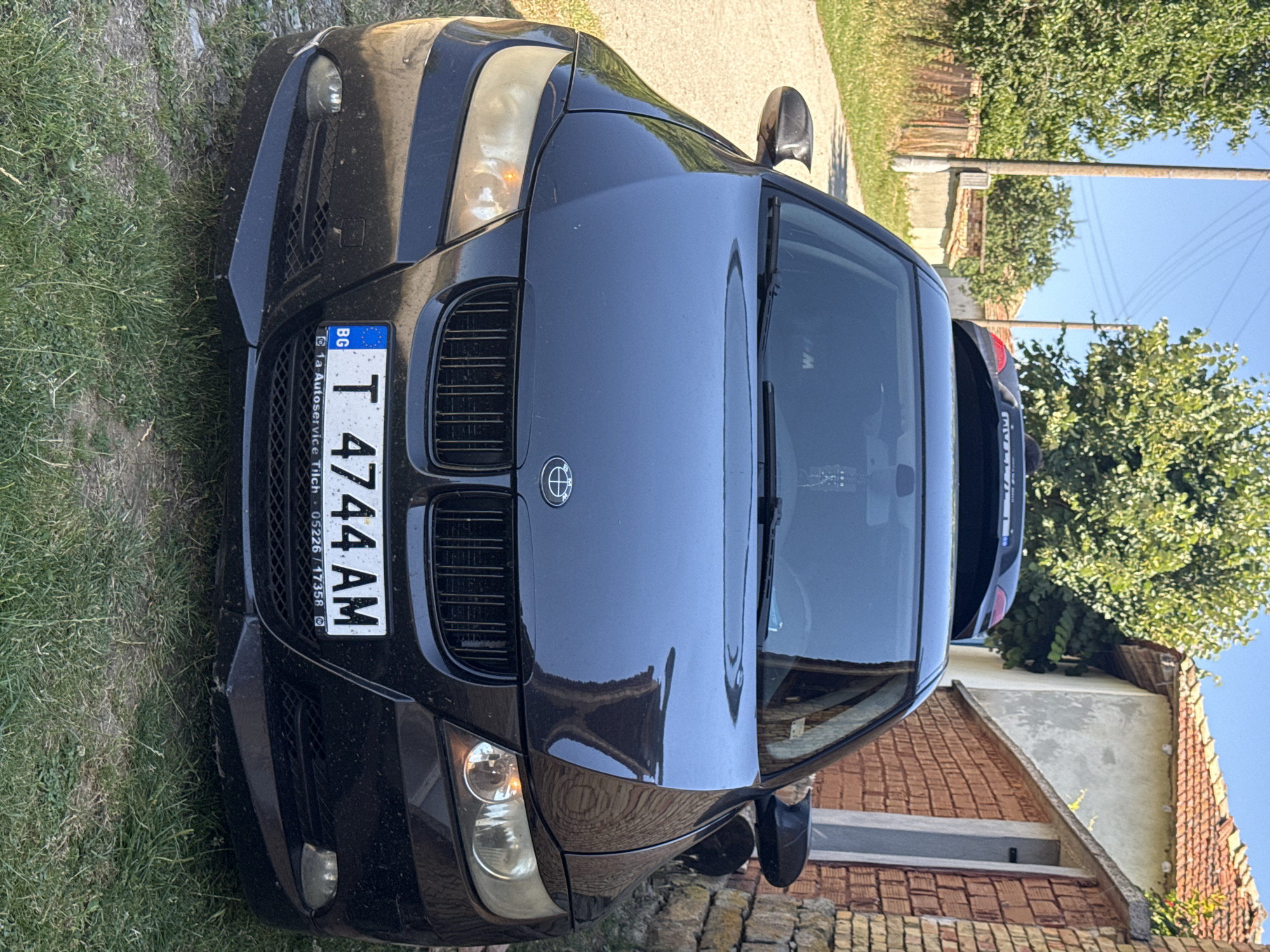 A black BMW sedan is parked on a grassy area next to a dirt road. The car faces the camera. The license plate reads "T 4744 AM" and has "BG" on the left, indicating Bulgaria. The car's headlights are on. Behind the BMW, another car, a dark color, is partially visible. The background shows a brick building with a tiled roof, trees, and a wooden fence. The environment appears to be rural. The image was taken in the city of Konop, Bulgaria.