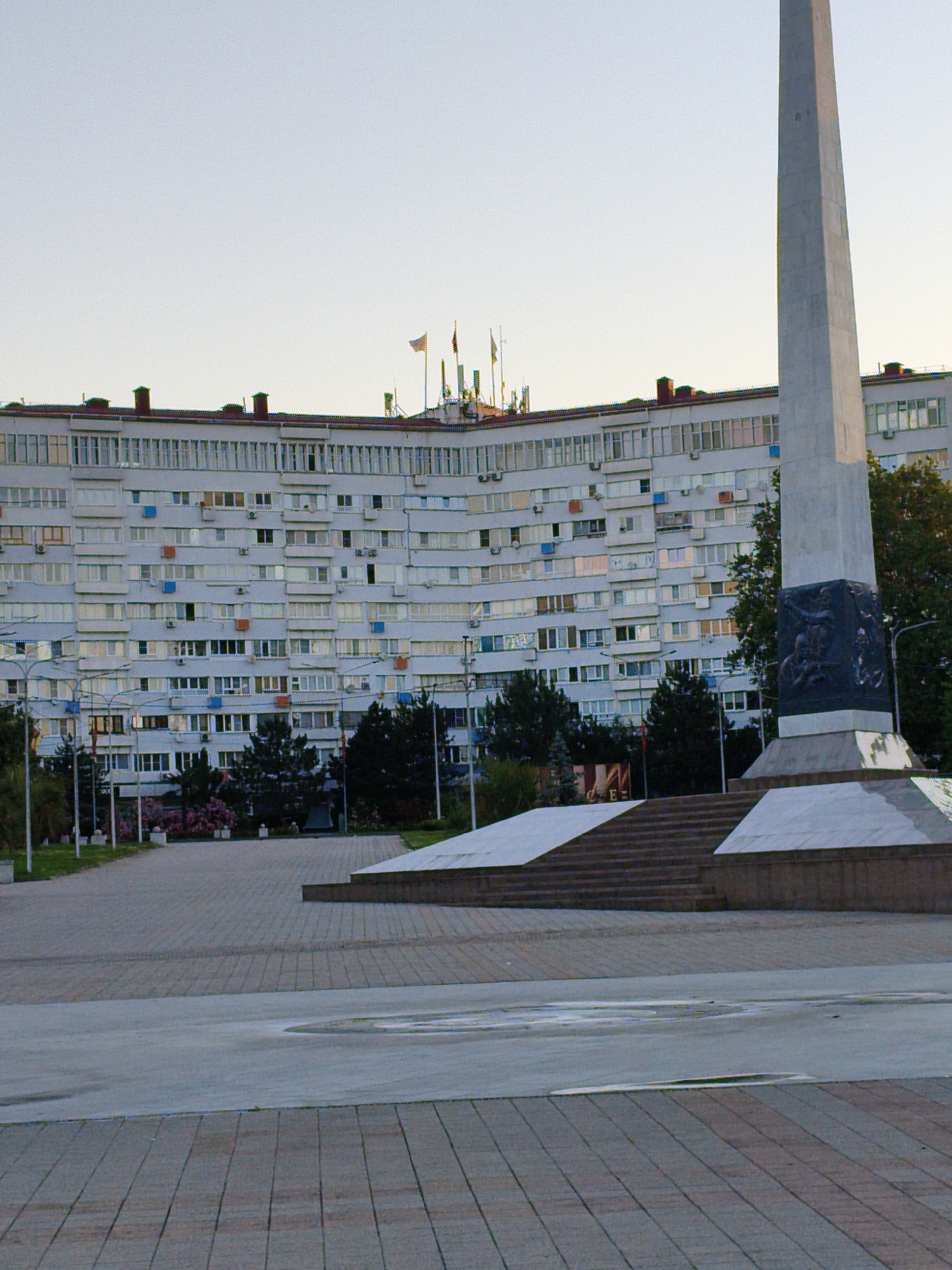 The image depicts a large, curved, multi-story apartment building with numerous windows, balconies, and visible air conditioning units. In the foreground, a tall, gray obelisk monument stands prominently, adorned with a dark, sculpted relief at its base. The monument is surrounded by a paved plaza with tiered steps and angled platforms. Trees and some flowering bushes are visible in the mid-ground, between the monument and the building. The sky above is clear and light. The scene appears to be an urban public space, likely a monument or memorial square within the city of Agoy, Russia, given the prompt's context. There are no people visible in the image.