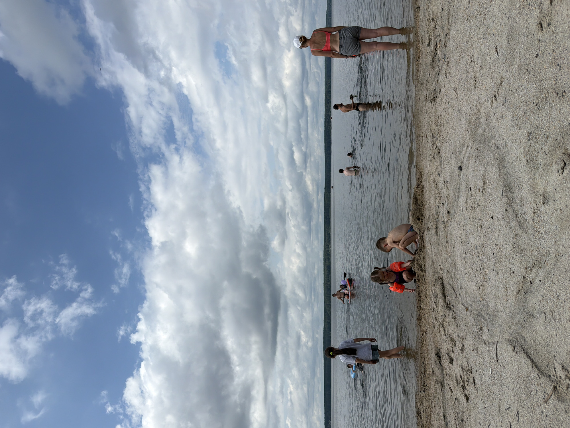 The image depicts a beach scene on a cloudy day, likely in the late afternoon or early evening given the diffused light. The sky is a mix of blue and heavy gray clouds, suggesting potential for rain. The setting appears to be a lake or large body of freshwater, with a sandy shoreline in the foreground.
Several people are present, engaged in various activities. Closest to the camera, two children are playing on the sand and in the shallow water. One child, wearing orange floaties, is crouching and appears to be digging or playing with the sand. Another child, a boy, is also crouching nearby. Further out in the water, several other individuals are visible. Some are standing or wading, while others are on paddleboards. One person is visible further in the distance, appearing to be swimming or standing in the water. The activity seems to be recreational, with people enjoying a day at the beach.
The overall mood of the image is relaxed and summery, despite the cloudy weather. There are no visible signs or text in the image. The location cues, such as the sandy beach and calm water, are consistent with a lake or a large river in Zarechny, Russia.