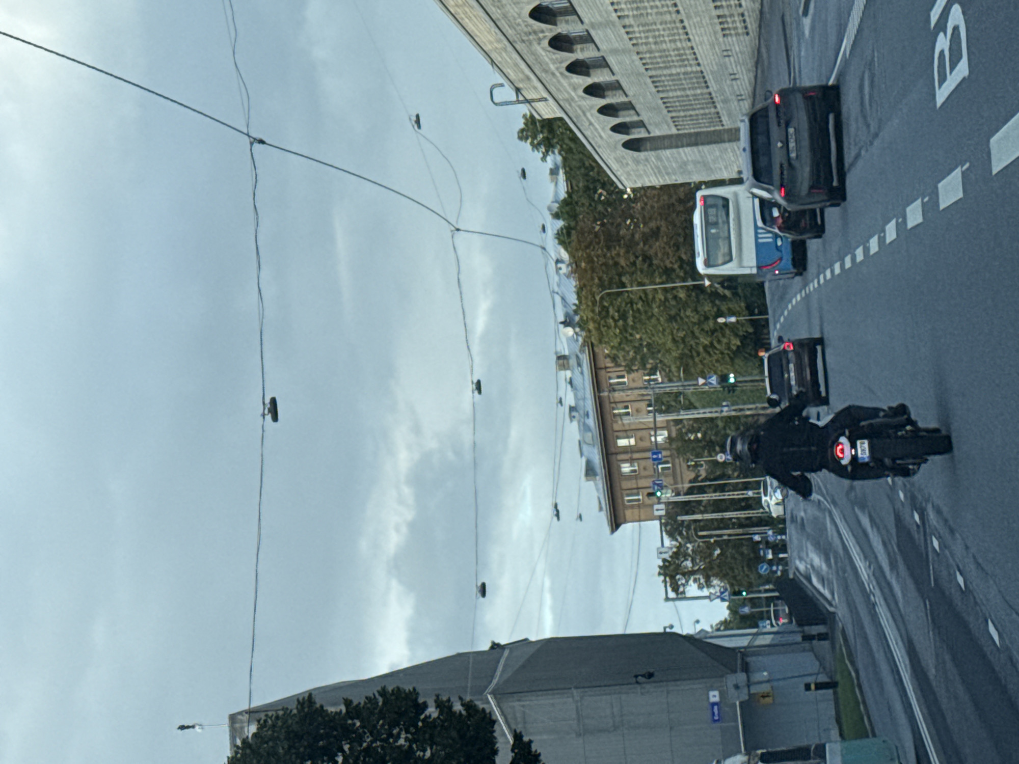 A motorcycle rider in a black outfit is seen from behind, riding on a wet asphalt road in Tallinn, Estonia. The rider is in the foreground, heading away from the viewer. The road has lane markings, and several cars and a bus are also on the road, in front of and around the motorcycle. The background shows buildings, trees, and utility wires crisscrossing the sky. The sky is overcast, suggesting a cloudy day. There are shoes hanging from some of the overhead wires.