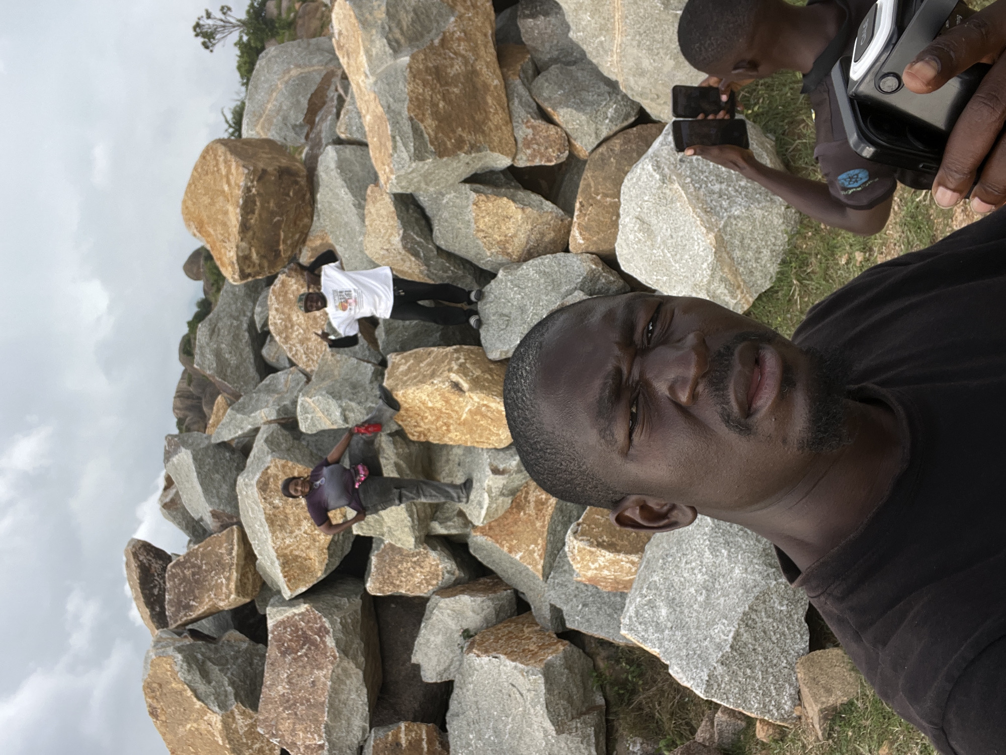 The image shows a group of people in a rocky outdoor environment. In the foreground, a man in a black t-shirt is looking at the camera, holding a black video camera. Behind him, another young person in a black t-shirt is holding two smartphones. Further in the background, a woman in a purple and grey outfit is posing on a large rock, and a man in a white t-shirt and black pants is standing on another rock, pointing his fingers upwards. The scene is composed of large, weathered rocks of various sizes and colors, set against a cloudy sky. This appears to be a natural rock formation, likely a tourist or recreational area. The presence of the video camera and smartphones suggests the individuals are documenting their experience. This location resembles the granite rock formations found in and around the city of Jos, Nigeria, known for their geological significance and scenic beauty.