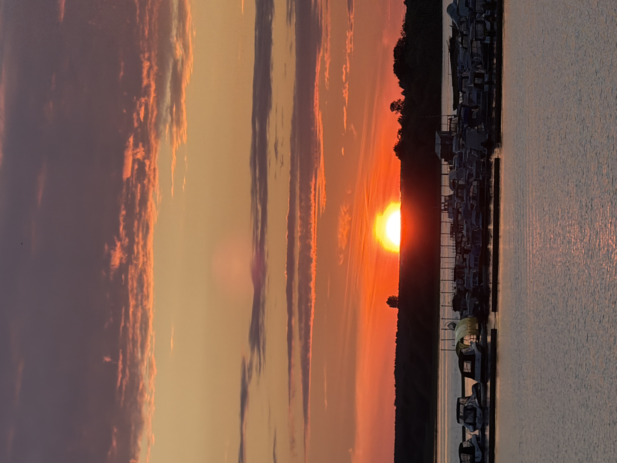 This is a vertical, landscape-oriented photograph taken at sunset. The sun, a bright yellow orb, is low on the horizon, casting a warm, orange and pink glow across the sky. The sky is filled with streaks of clouds, some illuminated by the setting sun in shades of orange and red, while others appear as dark, purplish stripes.
To the right of the frame, a dark silhouette of trees and vegetation forms a treeline along the shore. Adjacent to the treeline, a marina is visible, filled with numerous boats docked in rows. The boats appear to be of various sizes, mostly covered. The water in the foreground reflects the colors of the sunset, creating a shimmering, textured surface.
The scene evokes a sense of peace and tranquility, typical of a sunset over water. The colors are vibrant and saturated, indicating a clear or partly cloudy sky at dusk. There is no visible text in the image. The location cues, such as the marina and the type of vegetation, along with the provided context of Solikamsk, Russia, suggest this is a natural or recreational area near a body of water. The time of day is clearly sunset.