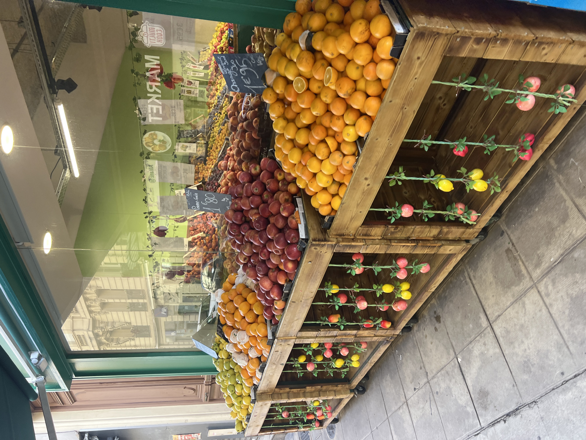 The image depicts an outdoor fruit stand located on a sidewalk in Nice, France, as indicated by a sign in the window displaying "MARKET" in red lettering and prices in Euros. The fruit stand features wooden crates filled with a variety of fresh fruits, including apples, oranges, and what appear to be plums and pears. The fruits are arranged in large piles, with some prices displayed on handwritten signs. Decorative artificial apples and lemons are hung on strings from the front of the wooden crates. The background shows the reflection of buildings in the storefront window, suggesting a street scene in a European city. The lighting appears to be natural daylight, supplemented by interior lighting from the shop. The ground is paved with gray square tiles. There are no people visible in the image.