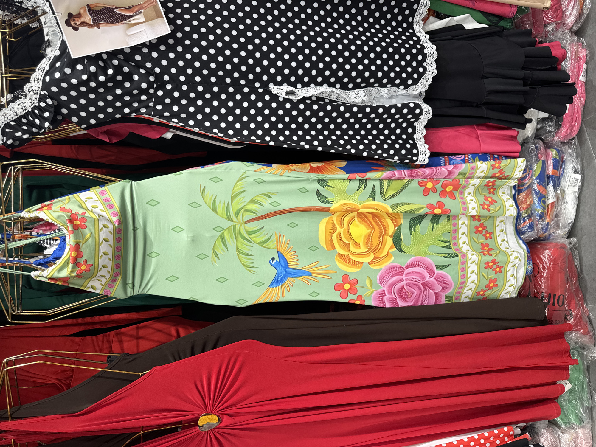 This image shows a rack of women's dresses for sale in a market in Gungoren, Turkey. The dresses are colorful and feature various patterns, including polka dots, floral prints, and tropical designs. The rack is made of gold-colored metal and the dresses are hung on hangers. There are stacks of bagged clothing items on the floor beneath the rack. The lighting is bright and the image is taken from a slightly low angle, focusing on the dresses.