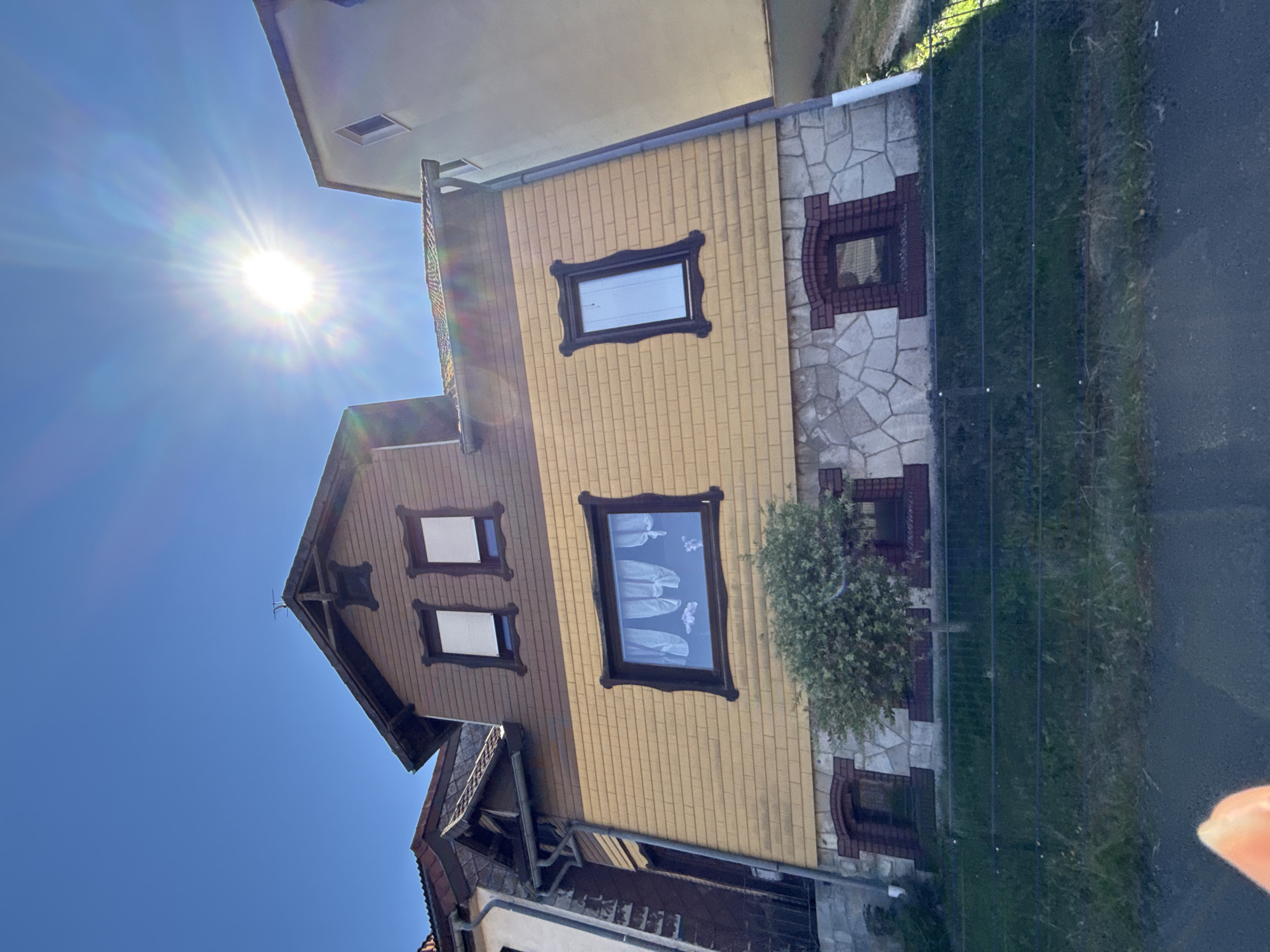 The image shows a multi-story house with a yellow shingled upper facade and a stone-clad lower facade. The house has several windows, some with white blinds and one with white curtains. A small tree stands in front of the lower facade. The house is situated in a sloped area with a green lawn and a dark fence in the foreground. A bright sun with lens flare is visible in the blue sky. The architecture and street view suggest this location is in the town of Bad Sachsa, Germany, specifically on a residential street.