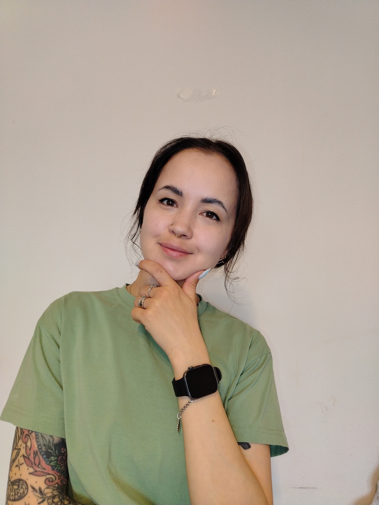 This is a portrait of a young woman taken indoors against a plain, light-colored wall, possibly in Noviy, Russia. She has dark hair, dark eyes, and is looking directly at the camera with a subtle, pleasant smile. Her right hand, adorned with several rings on multiple fingers and light blue nail polish on her thumb, is gently resting on her chin, suggesting a thoughtful or relaxed pose. She is wearing a simple, short-sleeved light green t-shirt. A black smartwatch with a matching strap is visible on her left wrist, which also reveals a sleeve of colorful tattoos on her forearm. She has two facial piercings: a small stud above a gold-colored hoop nose ring. The background wall is largely unadorned, though some minor imperfections or textures are visible in the upper portion. No text is visible in the image.