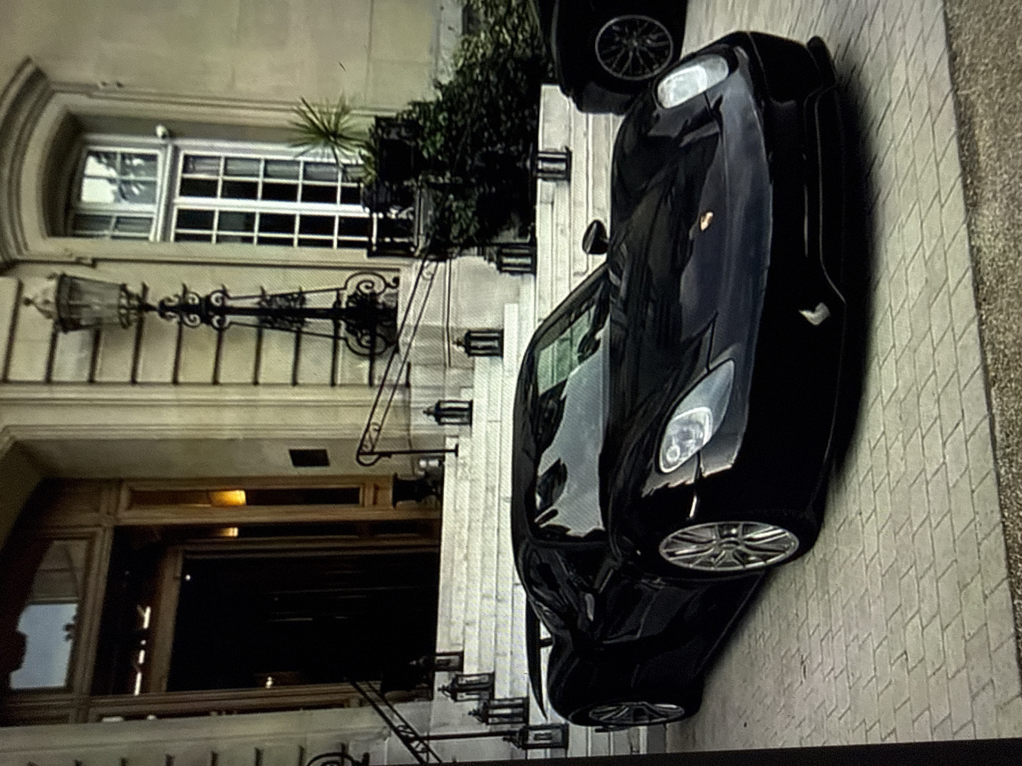 A black Porsche 918 Spyder is parked on a light-colored paver driveway in Kardzhali, Bulgaria. The vehicle is positioned in front of a grand, multi-story building featuring a light-toned facade and classical architectural elements. A wide, white stone staircase with black wrought-iron handrails leads up to a large, dark wooden double-door entrance. Several black metal lanterns are placed intermittently along the staircase. To the right of the entrance, an ornate black street lamp stands adjacent to a tall, arched window with multiple white panes. Green foliage and shrubs are visible at the base of the building and near the right side of the staircase. Partially visible on the far right, behind the Porsche 918 Spyder, is the front wheel and fender of another dark-colored vehicle with multi-spoke rims. The ground beneath the vehicles consists of rectangular pavers laid in a herringbone pattern, with a narrow strip of gravel at the bottom edge of the frame. No individuals are present in the scene.