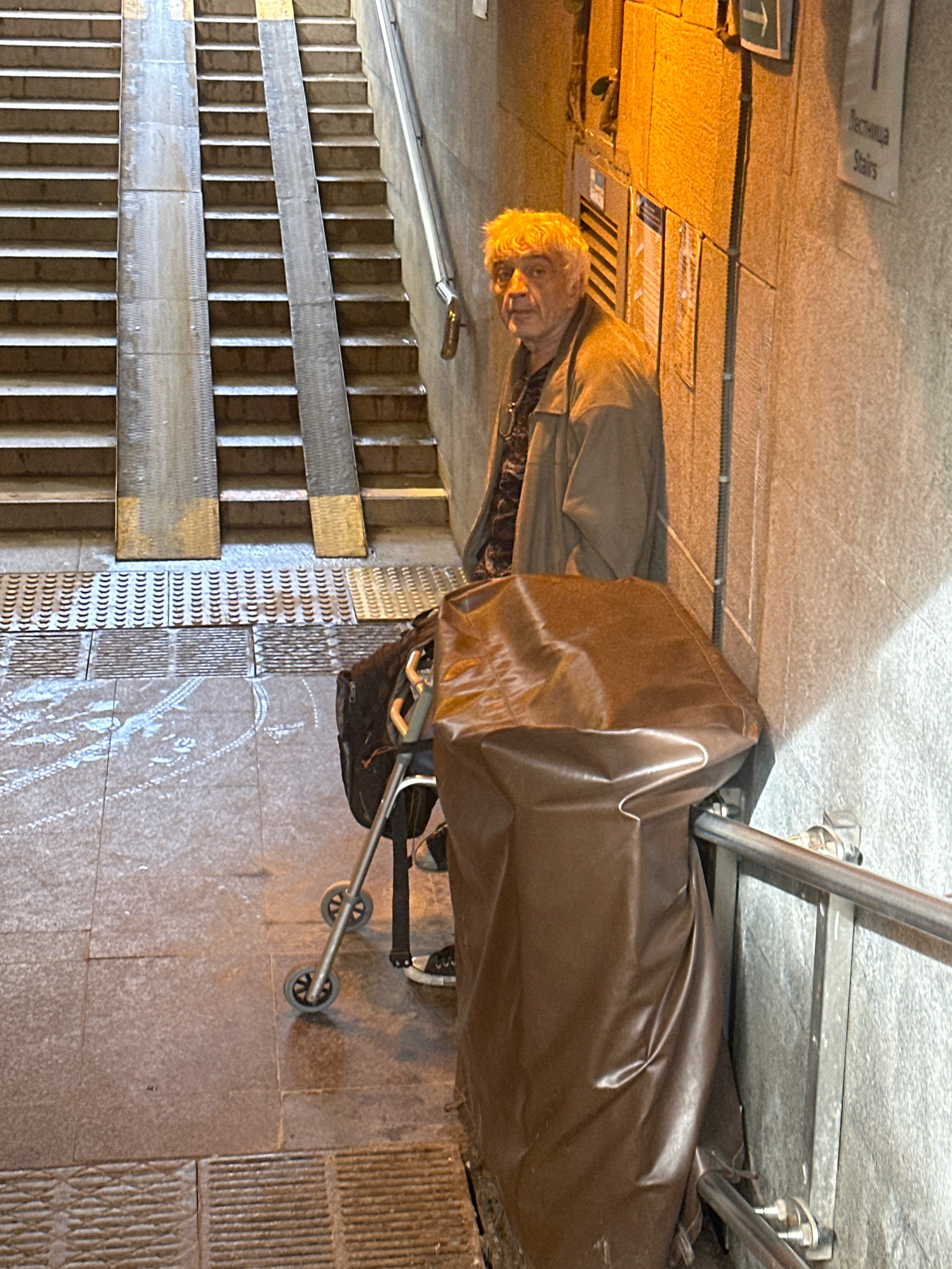 An elderly Caucasian male with white, curly hair stands in what appears to be an underpass or subway station in Vsevolozhsk, Russia. He is wearing a brown jacket over a patterned shirt and dark pants, and appears to be wearing dark shoes with white accents. He is leaning on a walker with two wheels, which has a black bag attached to it. In front of the walker and to the right of the man is a large object covered in brown, textured material, possibly a tarp or a cart. The man is looking towards the camera. The environment includes a concrete wall with signage that reads "Лестница Stairs" and an arrow pointing to the left. There is a set of stairs with metal ramps to the left of the man. The ground is paved with stone tiles, and there are some wet or reflective patches on the ground. The lighting is dim, with a strong yellow light illuminating the wall behind the man.